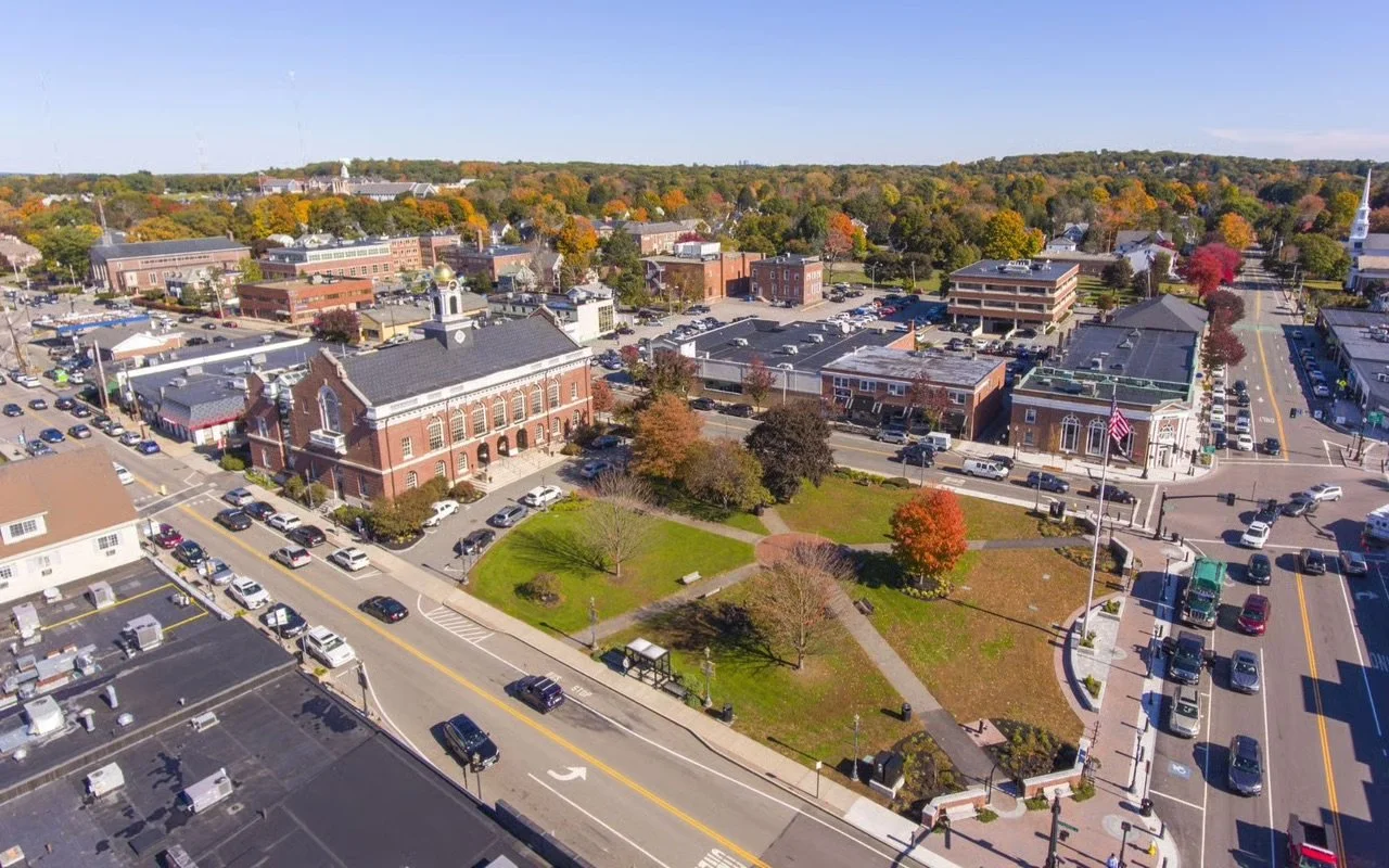 Aerial view of a small town square with historic buildings, a park with trees, and streets with parked and moving cars.