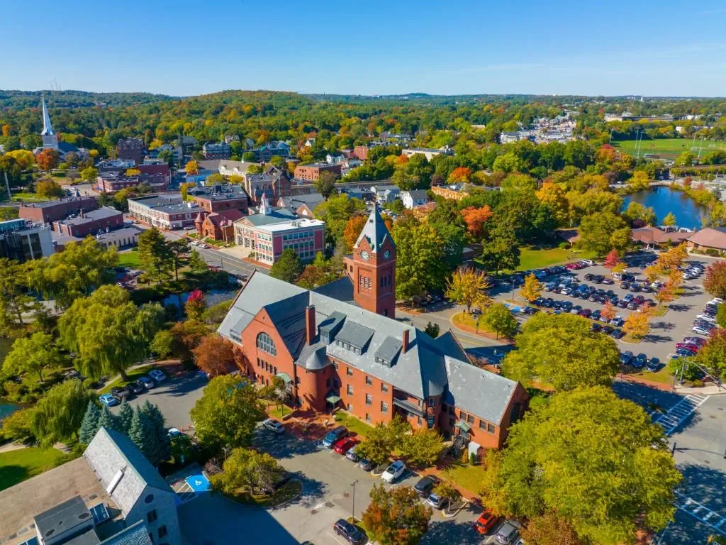 An aerial view of a small town with colorful trees, a large red brick building with a clock tower, parking lots, lakes, and a mix of historic and modern buildings under a clear blue sky.