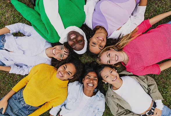 Six women of diverse ethnicities lying on grass in a circle, smiling at the camera.
