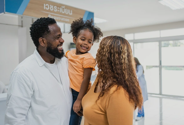 A smiling family of three at an airport, with a father, mother, and their young daughter.