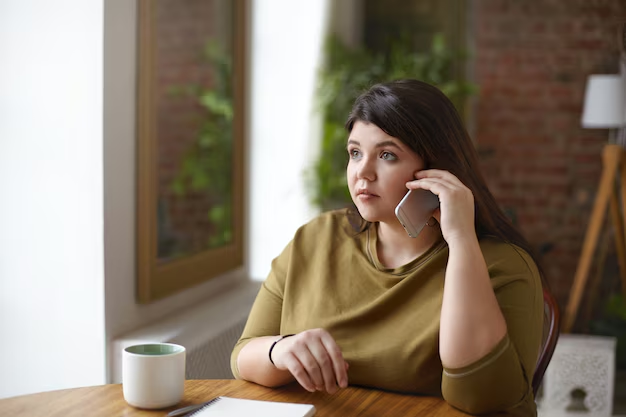 A woman with brown hair, wearing a mustard yellow shirt, sitting at a wooden table, talking on a phone, with a white coffee mug and a notepad in front of her. She is inside a room with large windows, a brick wall, and some greenery outside.