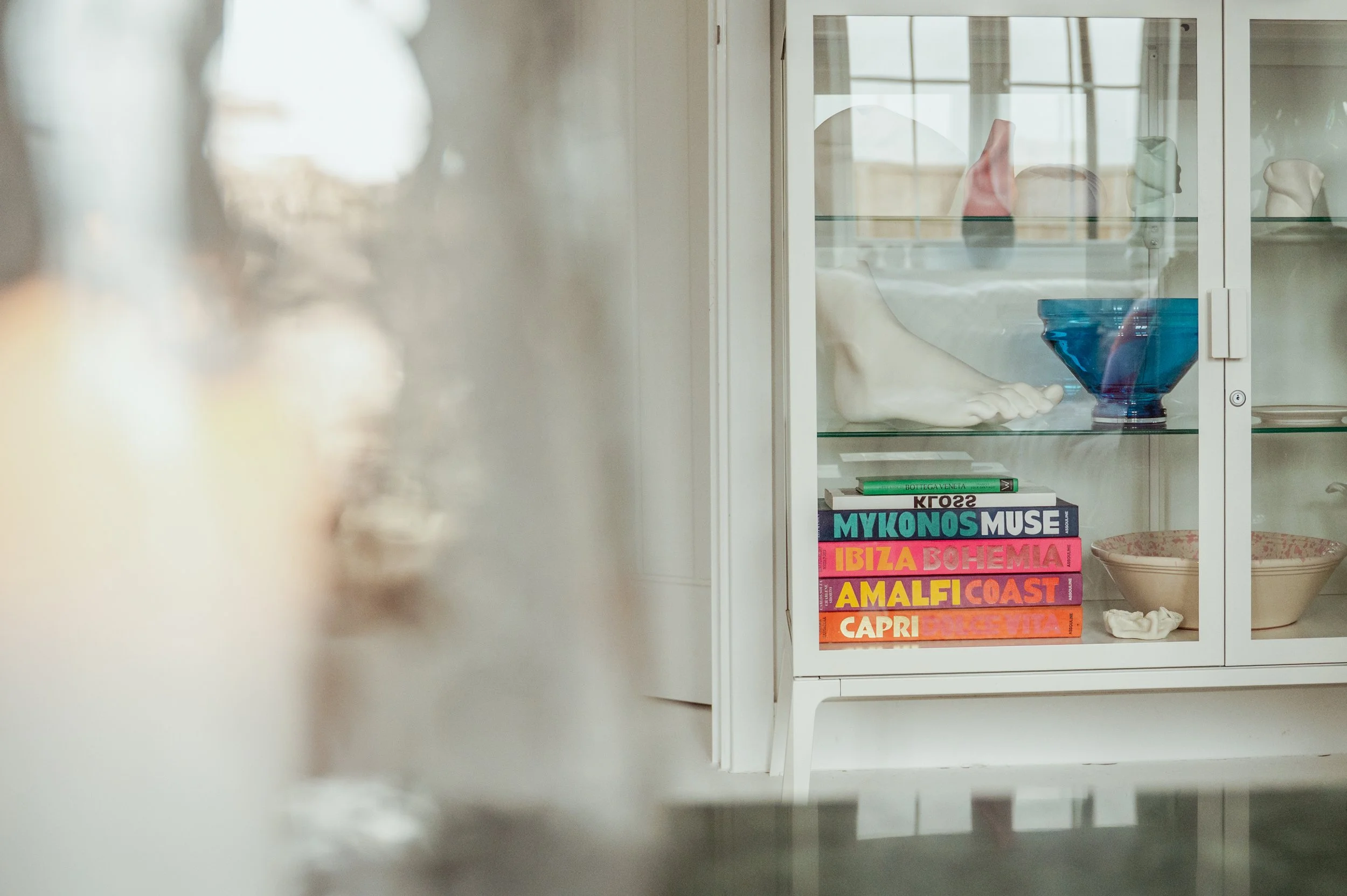 A white display cabinet with decorative objects including a white foot sculpture, colorful vases, and a stack of travel books about Greek islands and European destinations, with a beige bowl on the bottom shelf.