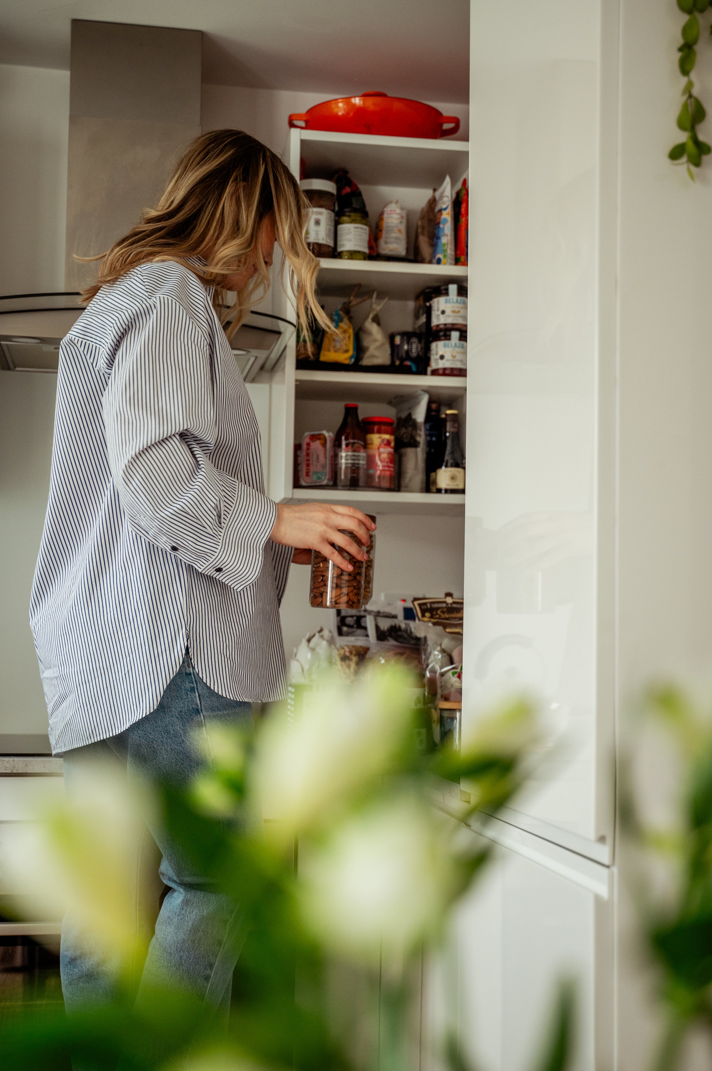 A woman with wavy blonde hair wearing a striped shirt and jeans is reaching into an open kitchen cabinet filled with jars, bags, and canned goods, with a blurred green plant in the foreground.