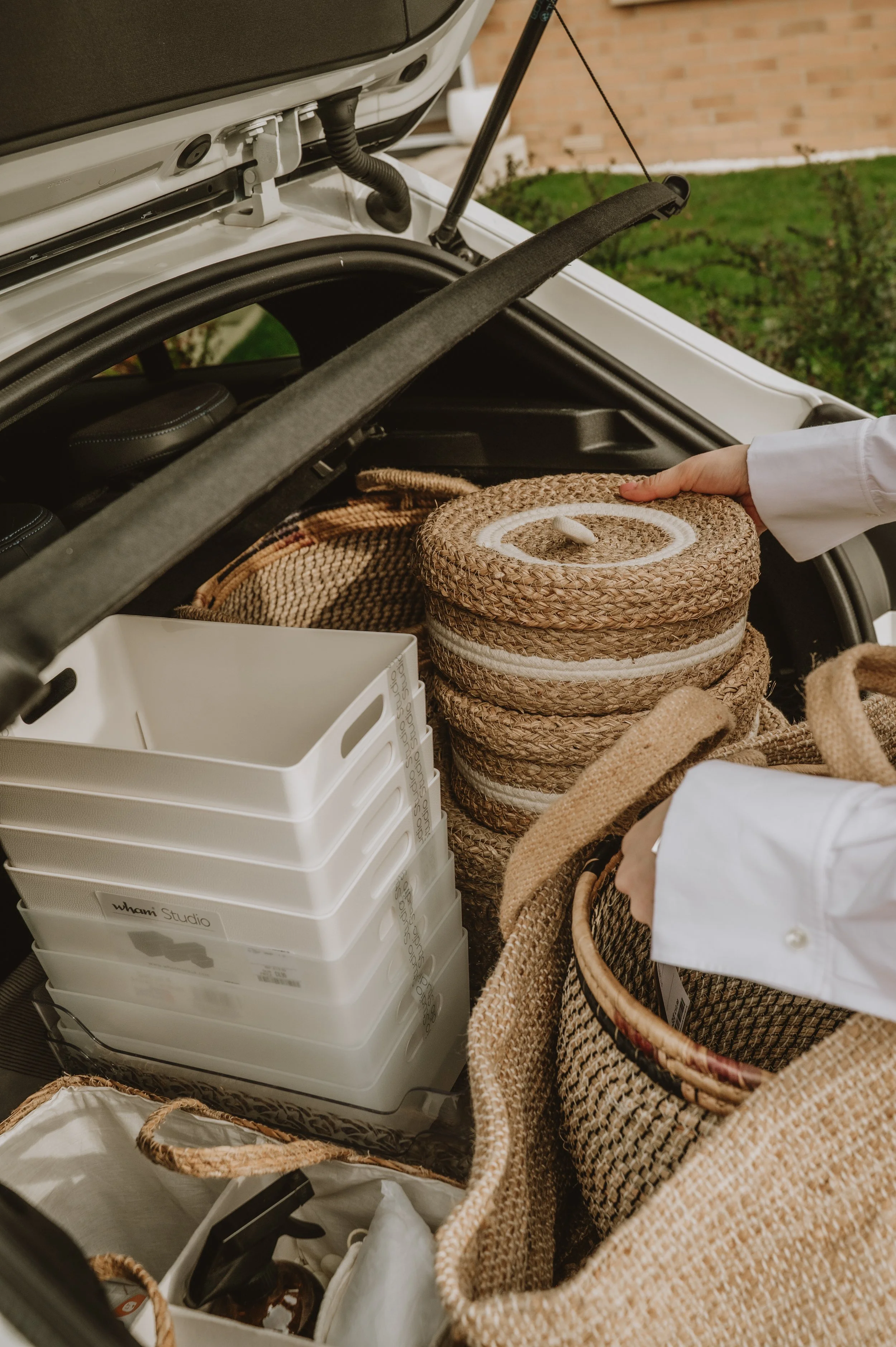 The open trunk of a car containing woven baskets, a white plastic storage bin, and a blanket, with a person's hand reaching inside.