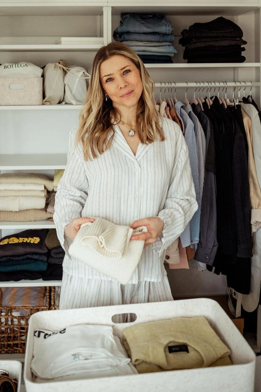Woman in striped pajamas standing in a closet, holding a folded cream-colored sweater, with shelves of folded clothes and hanging garments in the background.