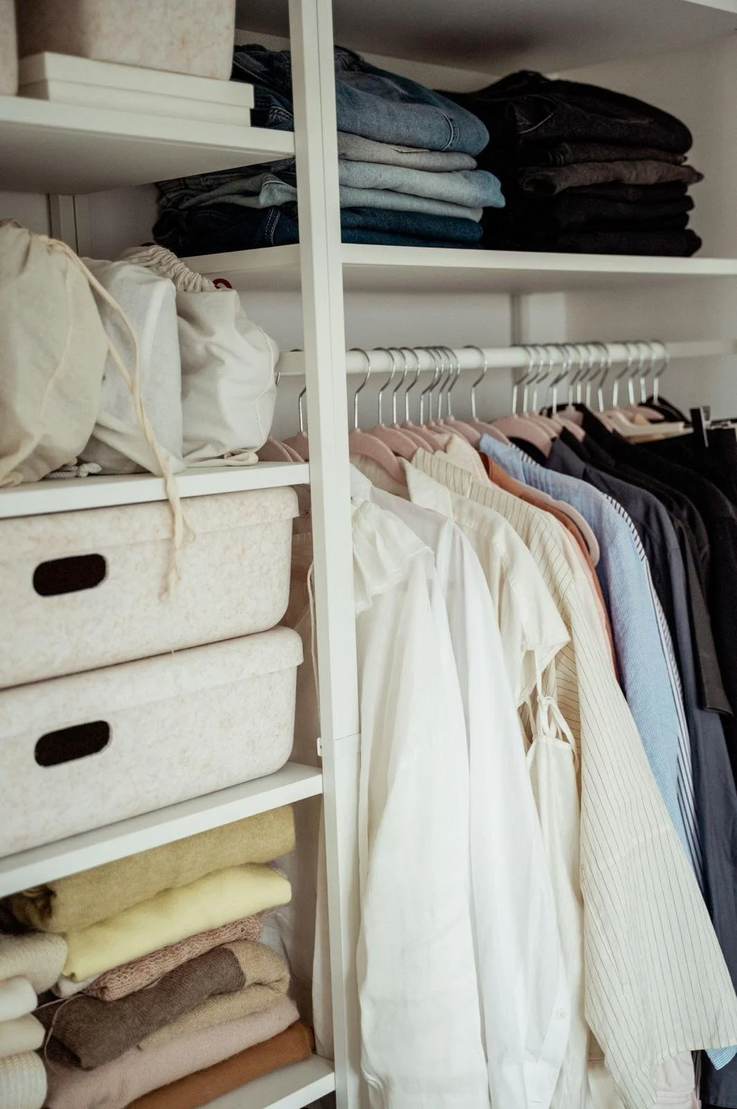 Closet with neatly folded jeans, stacked on shelves, and shirts hanging on a wardrobe rail, with storage bins and folded towels or linens on the shelves.