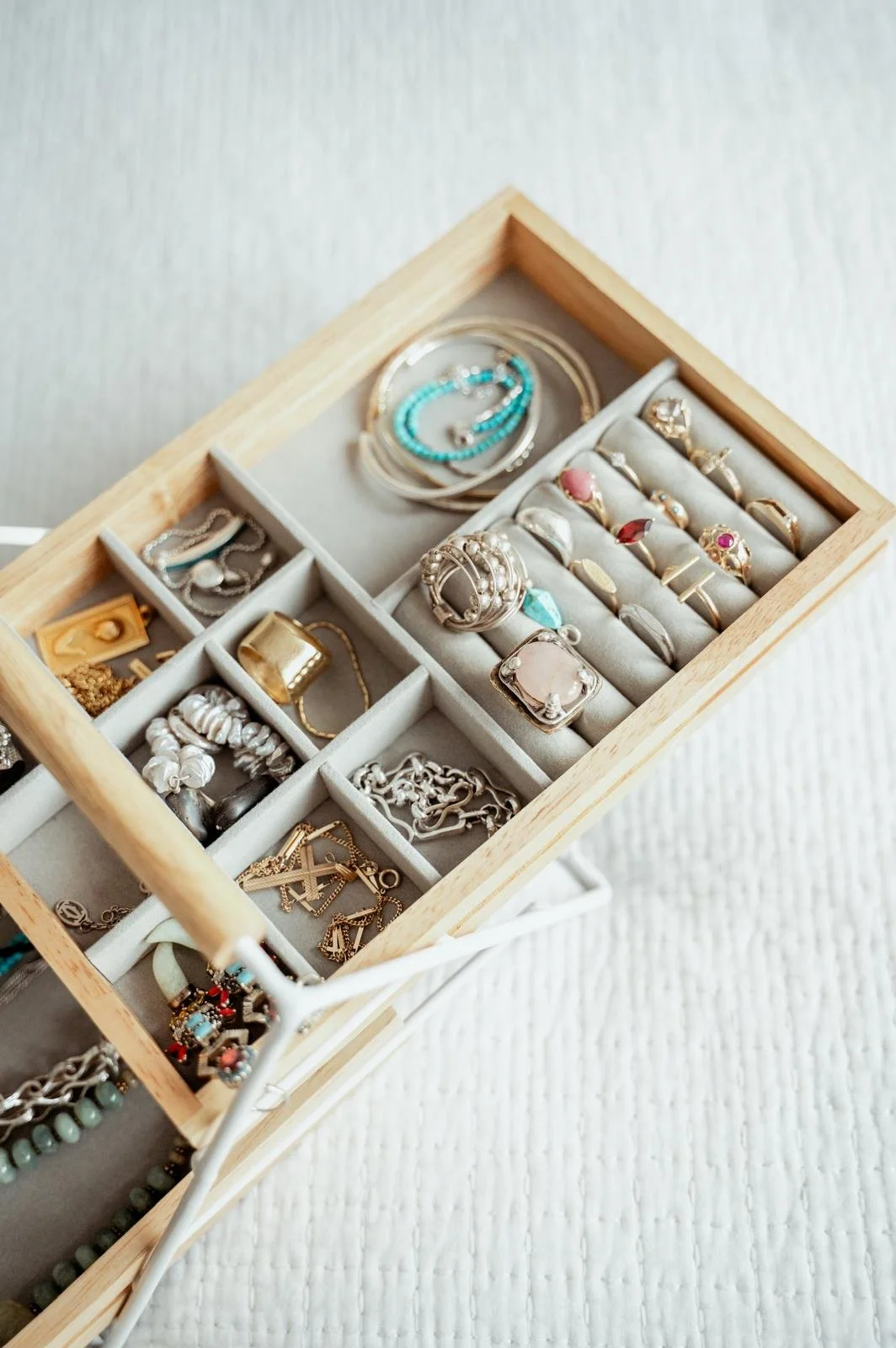 A wooden jewelry box with multiple compartments filled with rings, bracelets, and necklaces, displayed on a textured white surface.