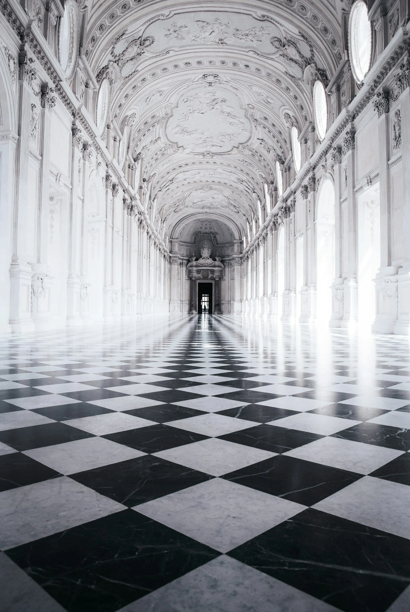 Interior of a grand, ornate hall with high, decorated ceilings and large windows, featuring a black and white checkered marble floor and a silhouette of a person near the far doorway.