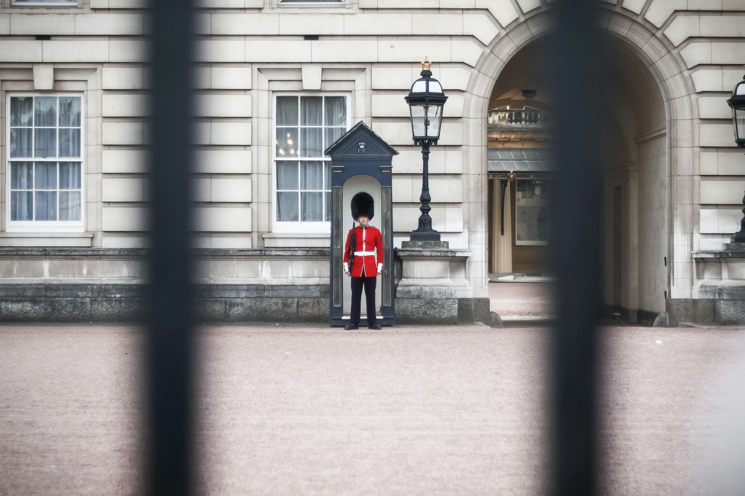 A Royal Guard in a red uniform and black tall hat standing outside a building, viewed through black iron bars.