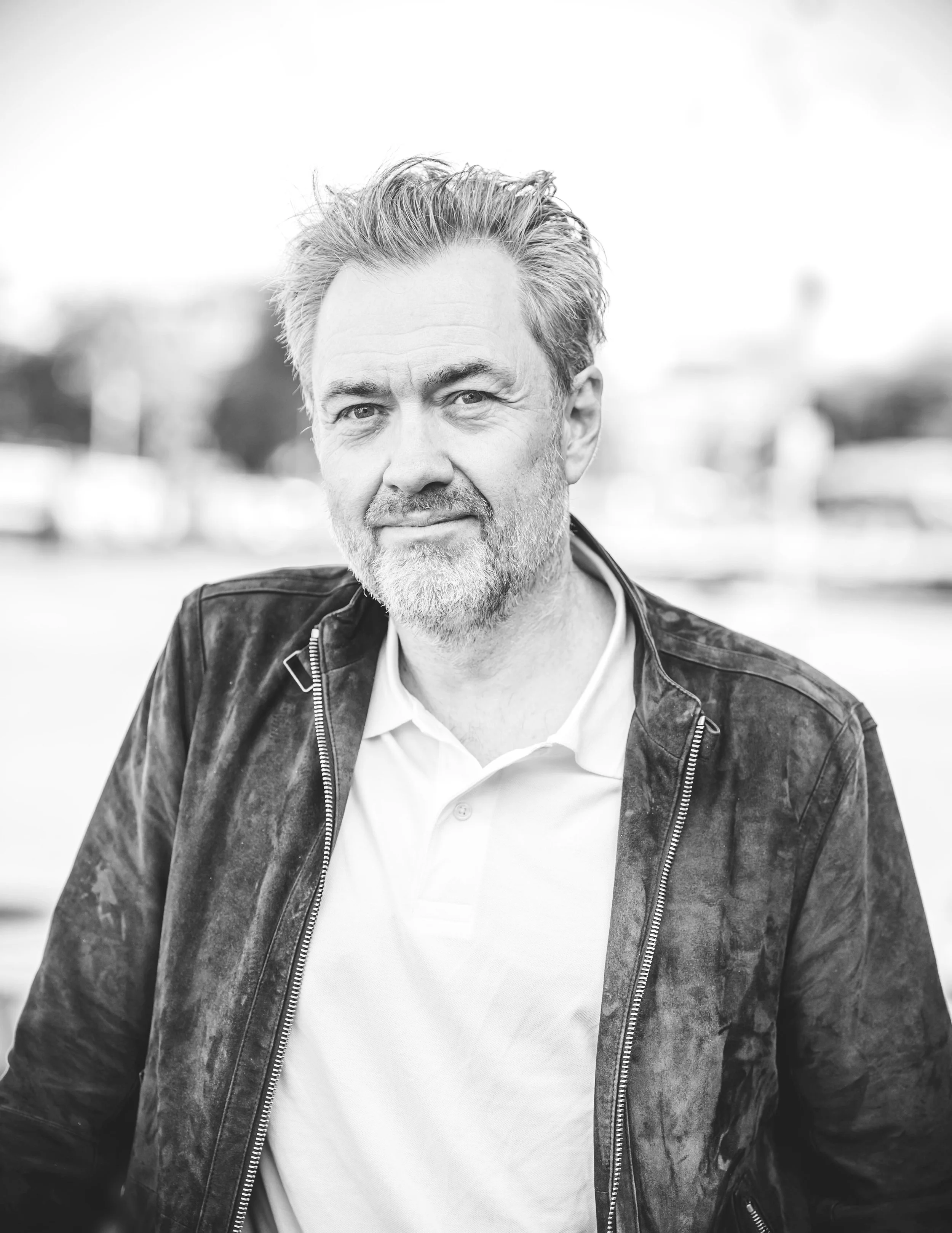Black and white portrait of a middle-aged man with gray hair and beard, wearing a casual jacket over a polo shirt, outdoors.