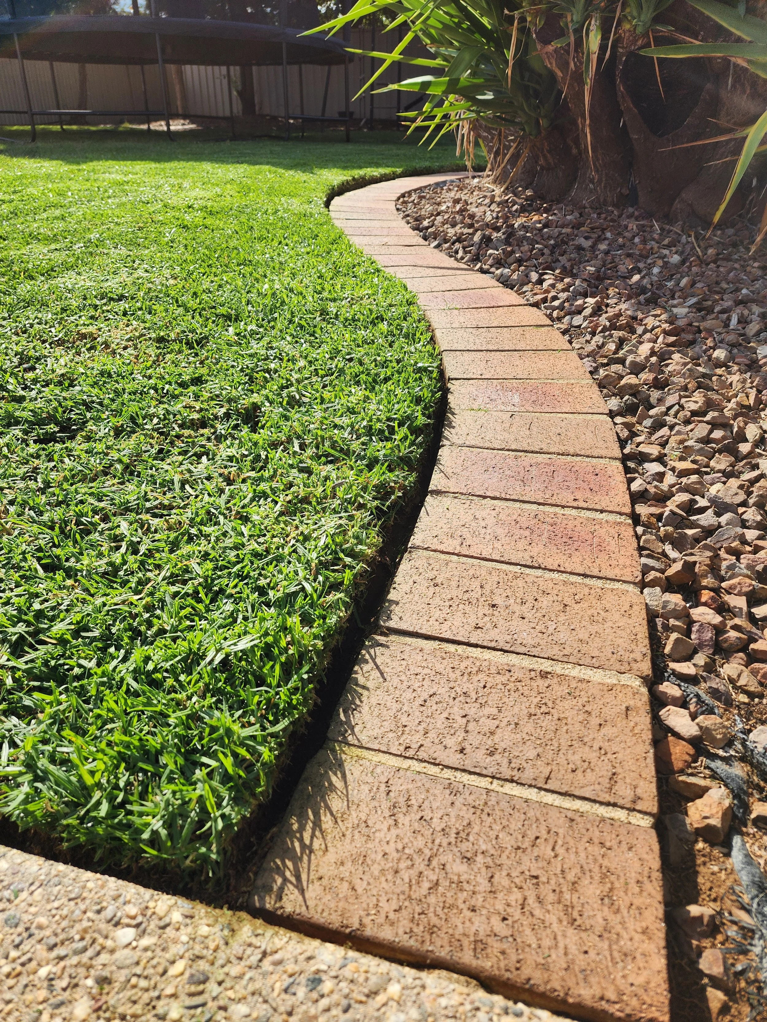 A curved brick pathway bordered by a lush green lawn on one side and a garden bed with rocks and plants on the other, with a metal fence and a trampoline visible in the background.