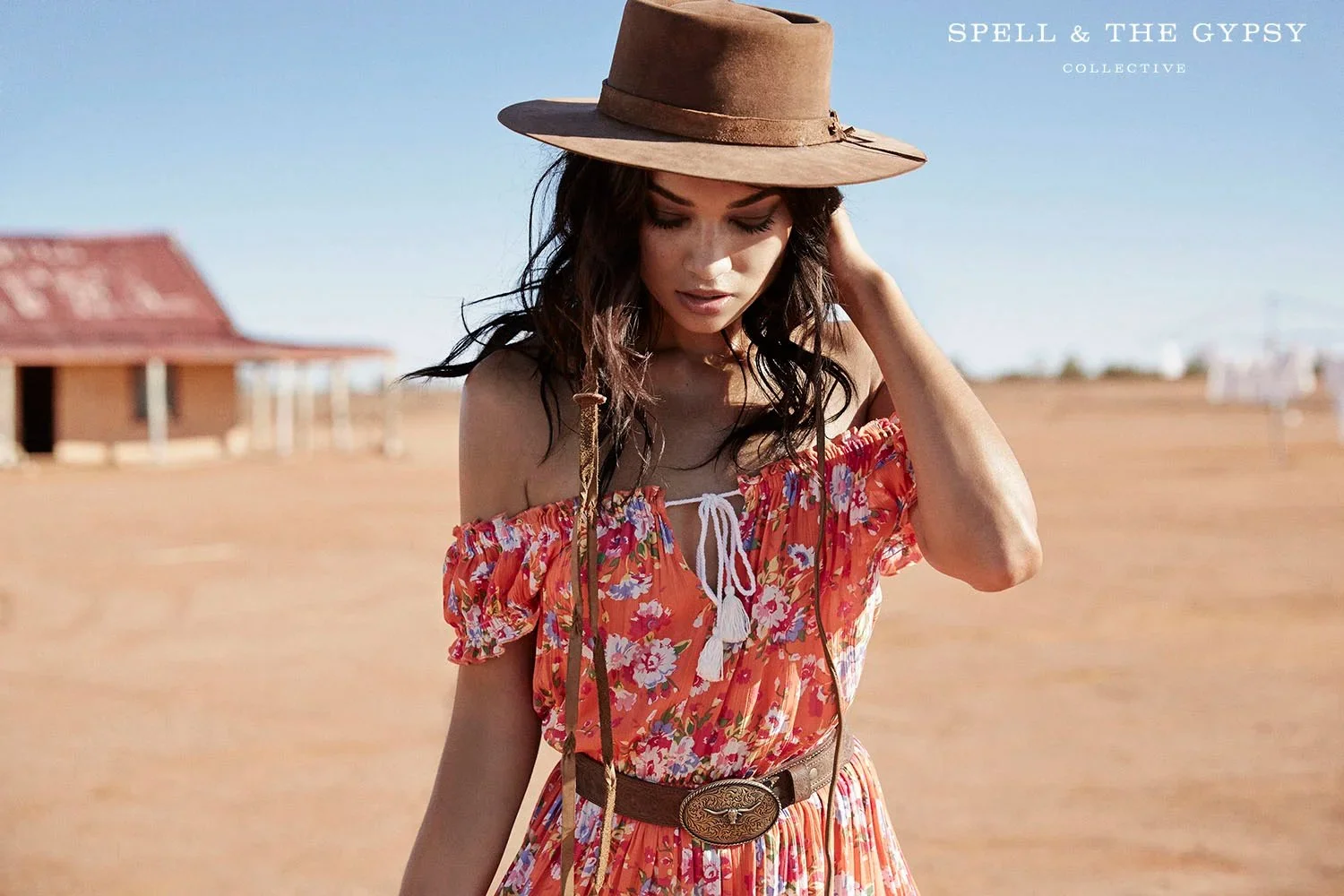 A woman in a floral off-shoulder dress with a wide-brimmed hat standing outdoors on a sunny day, with an expansive desert landscape and an old building in the background.