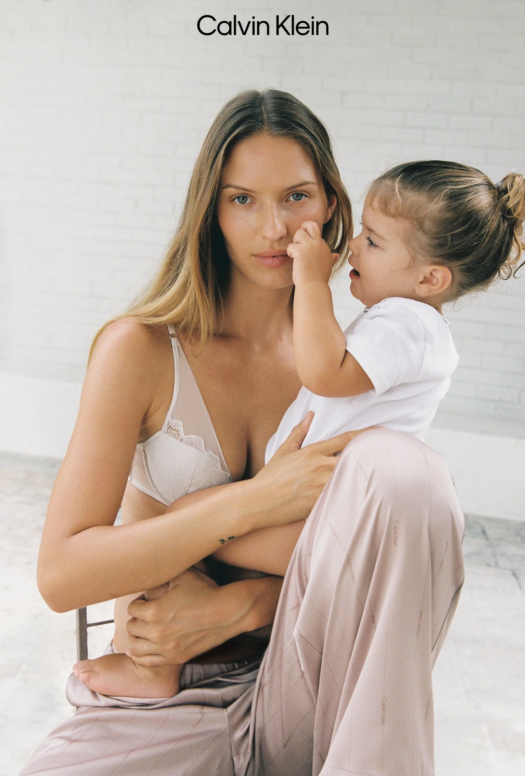 A woman with long blonde hair and a young girl with curly hair are sitting together against a white brick wall. The girl is touching the woman's nose, and the woman is looking directly at the camera. The woman is wearing a light-colored bra and loose