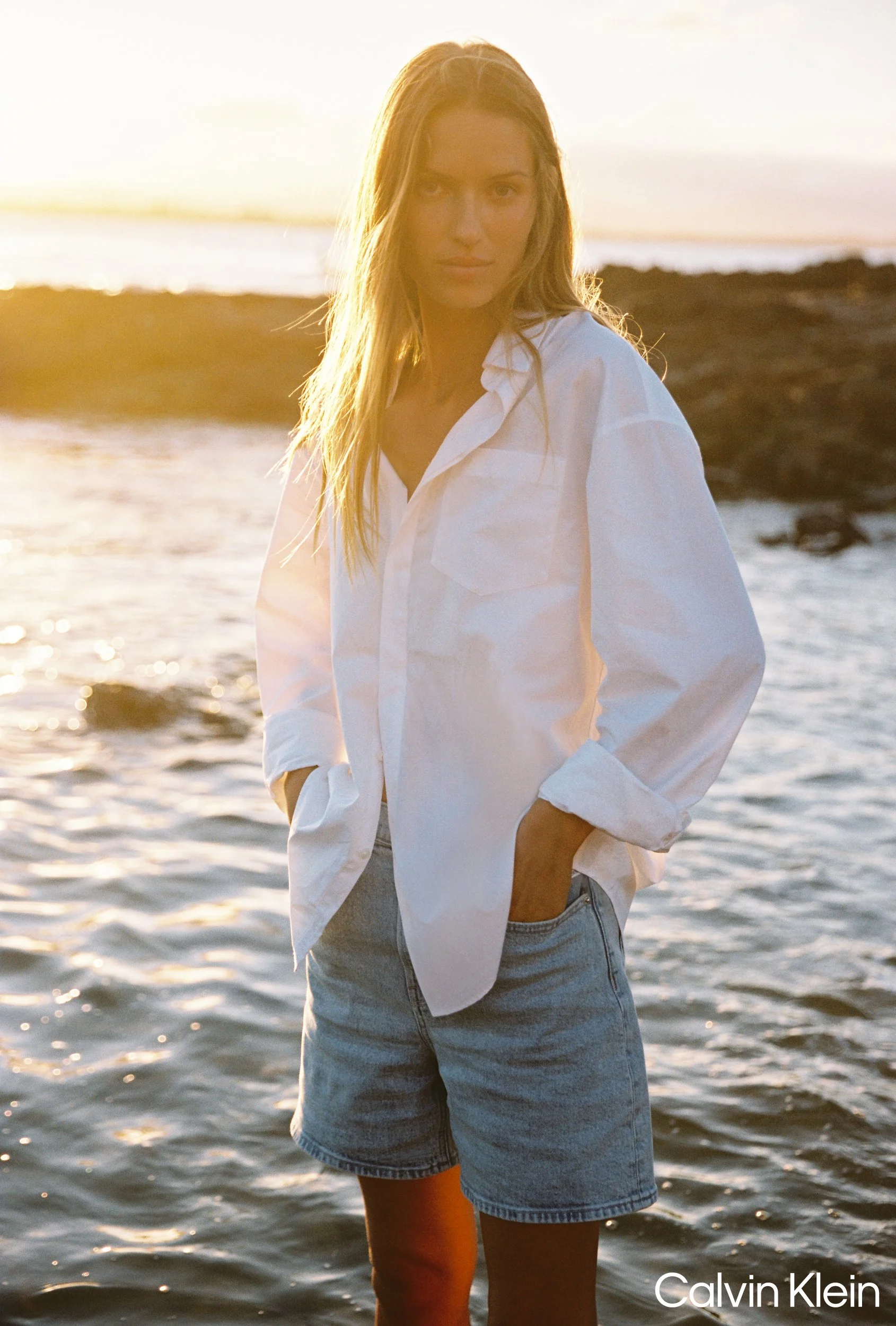 A woman standing in front of water during sunset, wearing a white shirt and denim shorts.