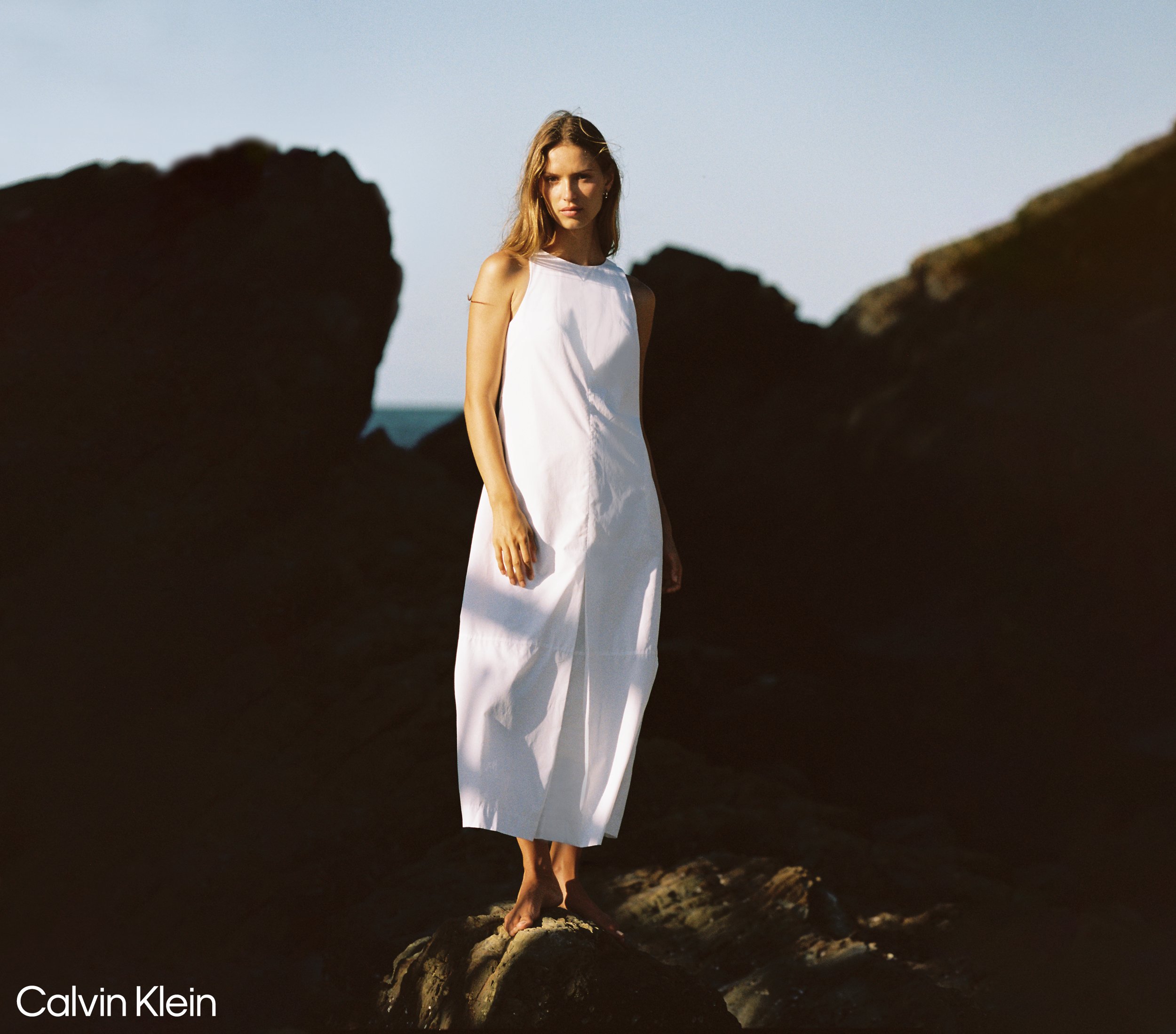 A woman standing on rocks at the beach, wearing a long white dress, with the sunset or sunrise lighting casting a warm glow.