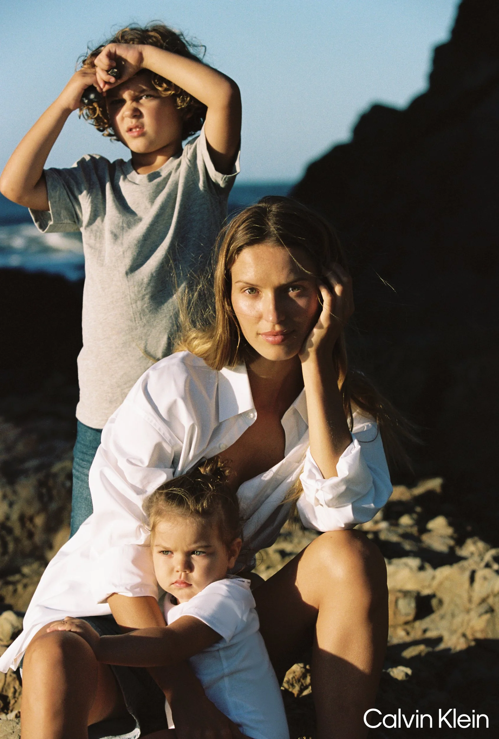 A woman with long brown hair sitting on rocks at the beach with two children, one hugging her and the other standing behind her shielding her eyes from the sun with his hand during sunset.