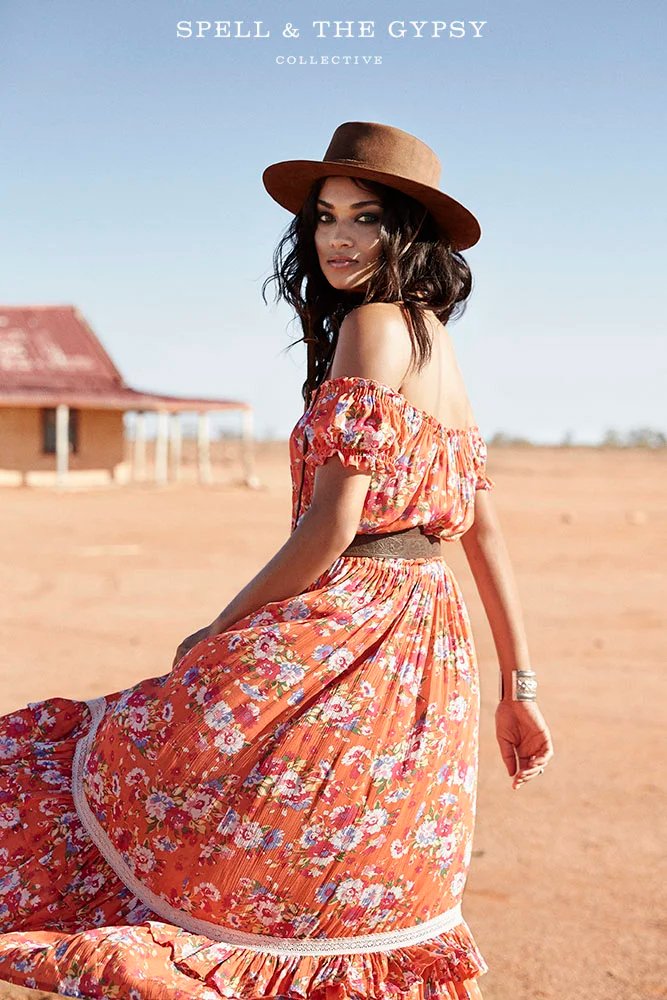 A woman in a floral off-shoulder dress and wide-brimmed hat sitting outdoors in a desert-like setting with a building in the background. She has dark hair and is wearing a bracelet.