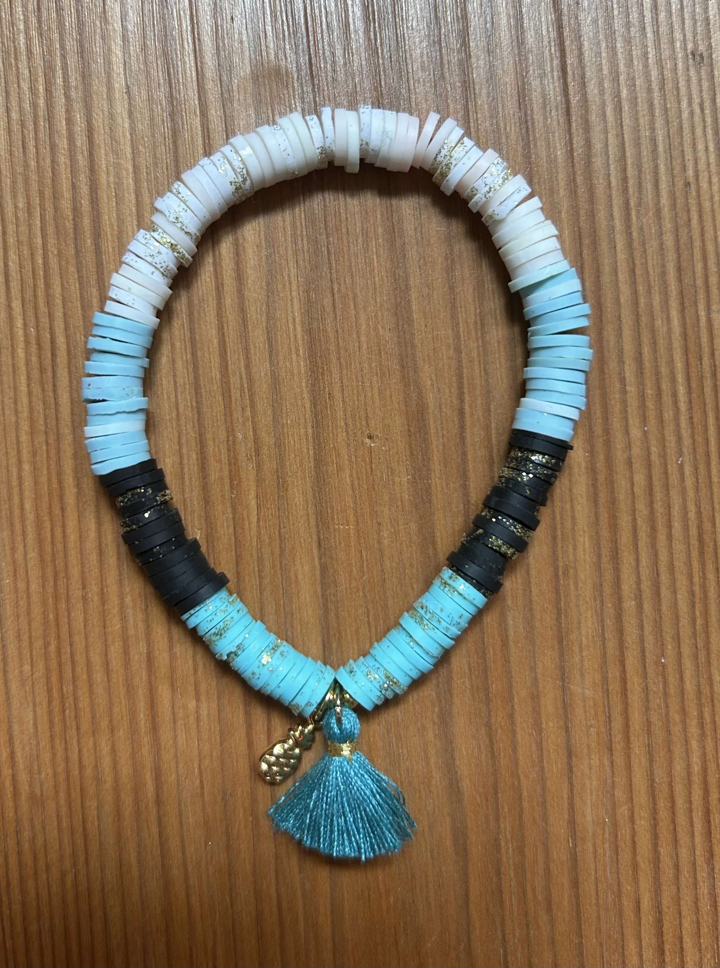Strandkette aus blauen, weißen, schwarzen, und durchsichtigen Scheiben mit einer Anhänger aus Gold und einer blauen Quaste, liegt auf einem Holzoberfläche.