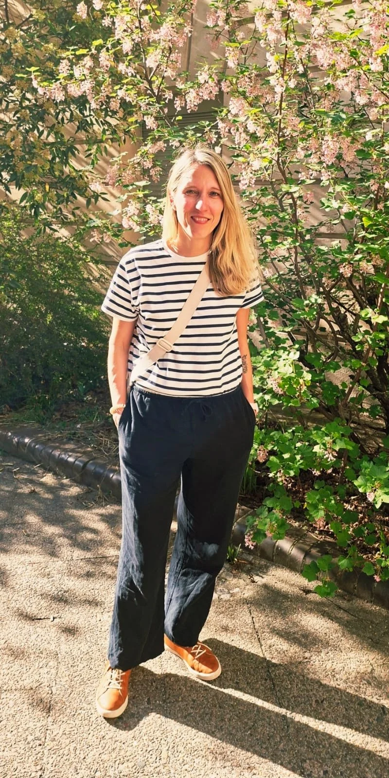 Frau mit langem blonden Haar, gestreiftem T-Shirt, schwarzen weiten Hosen, orange Schuhe, steht vor blühendem Strauch mit rosa Blüten, sonniger Tag, im Freien.