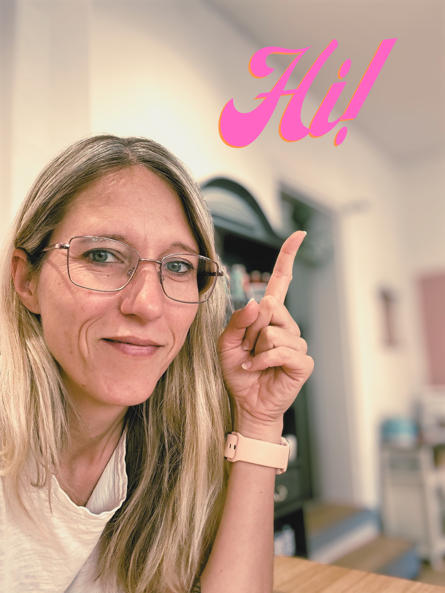 Ein Mann mit Brille und langen blonden Haaren, der auf seine Hand zeigt, auf der ein pinkfarbenes Textstempel mit dem Wort "Hi!" angebracht ist, in einem Raum mit Bücherregalen im Hintergrund.