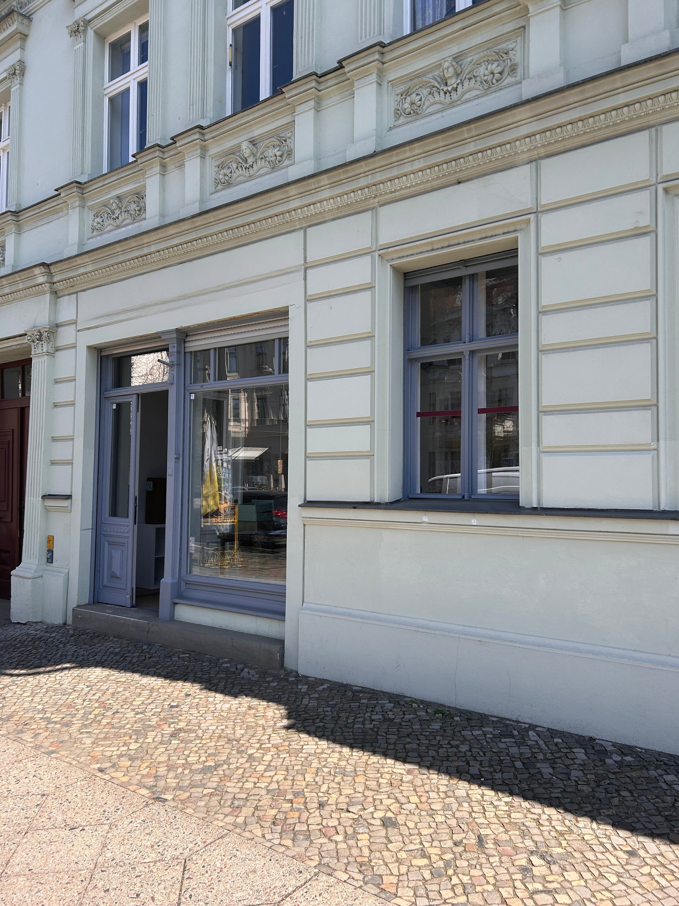 Einhelliges Bild eines Gebäudes mit weißen Fassaden und großen Fenstern. Das Erdgeschoss hat eine Tür und große Fenster mit bläulichem Rahmen. Der Gehweg besteht aus kleinen bunten Pflastersteinen, während der Schatten des Gebäudes auf den Gehweg fällt.