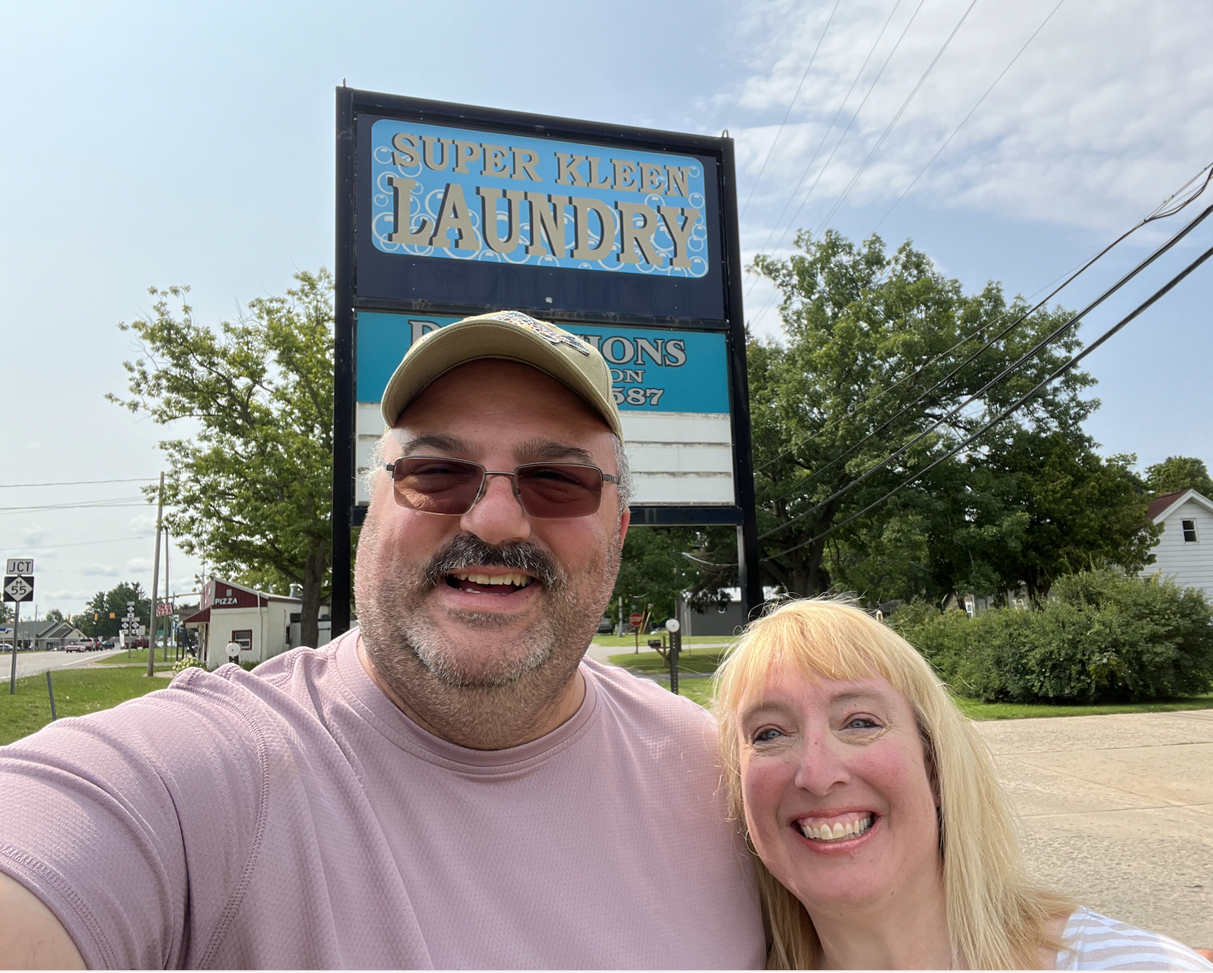 A smiling man in sunglasses and a woman with blonde hair taking a selfie outside of a laundry and dry cleaning business with a sign that reads 'Super Kleen Laundry' in the background.