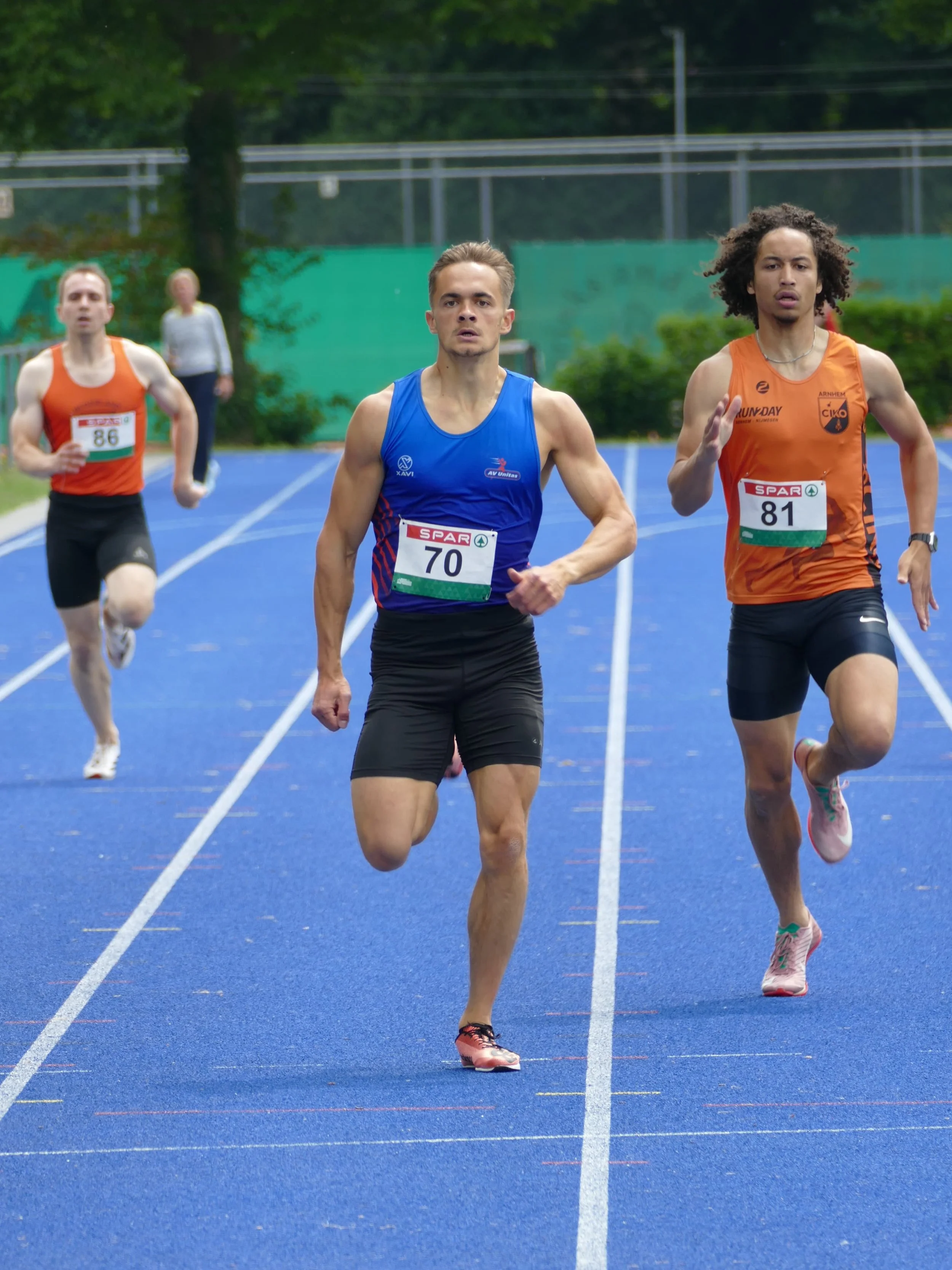 Drie atleten rennen op een blauwe atletiekbaan tijdens een race, met groene bomen en een hek op de achtergrond.