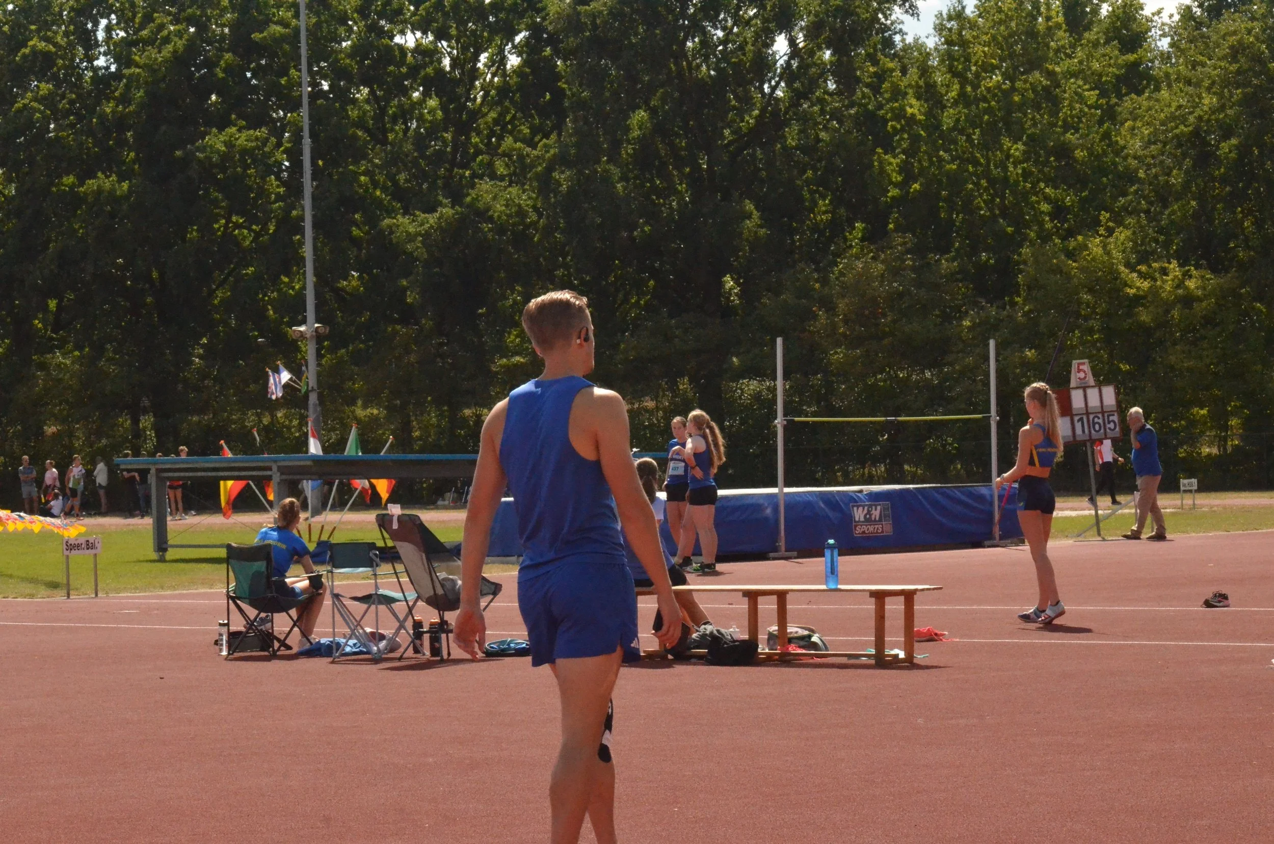 Atleet in blauw sportuniform die op een atletiekbaan staat, met meerdere mensen en sportstructuren op de achtergrond, onder een boomrijke hemel.