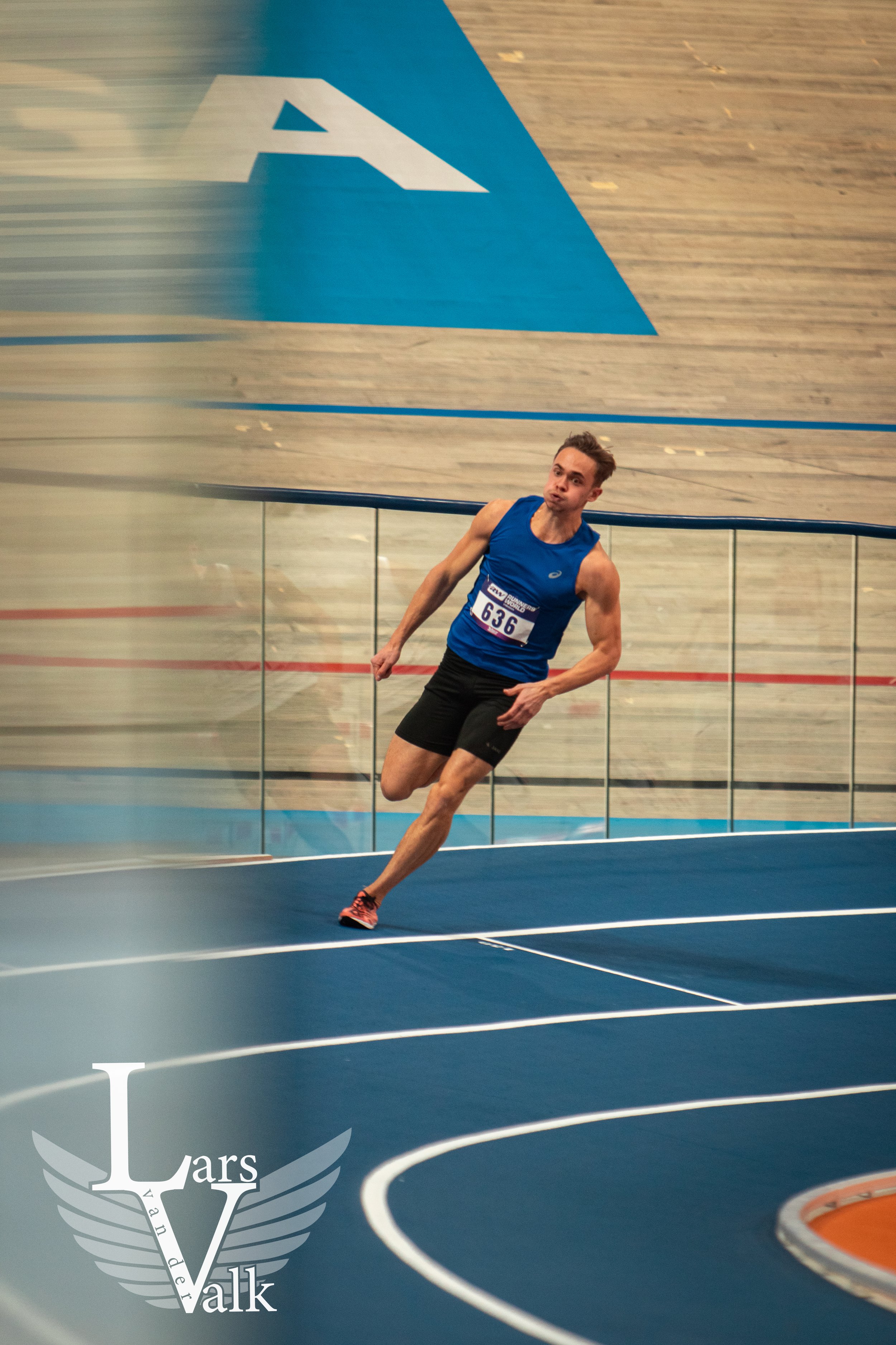 Een man rent in een indoor atletiekbaan terwijl hij '[636]' op zijn atletieknummer draagt, en er staat een logo 'Lars Vank' onderaan links.