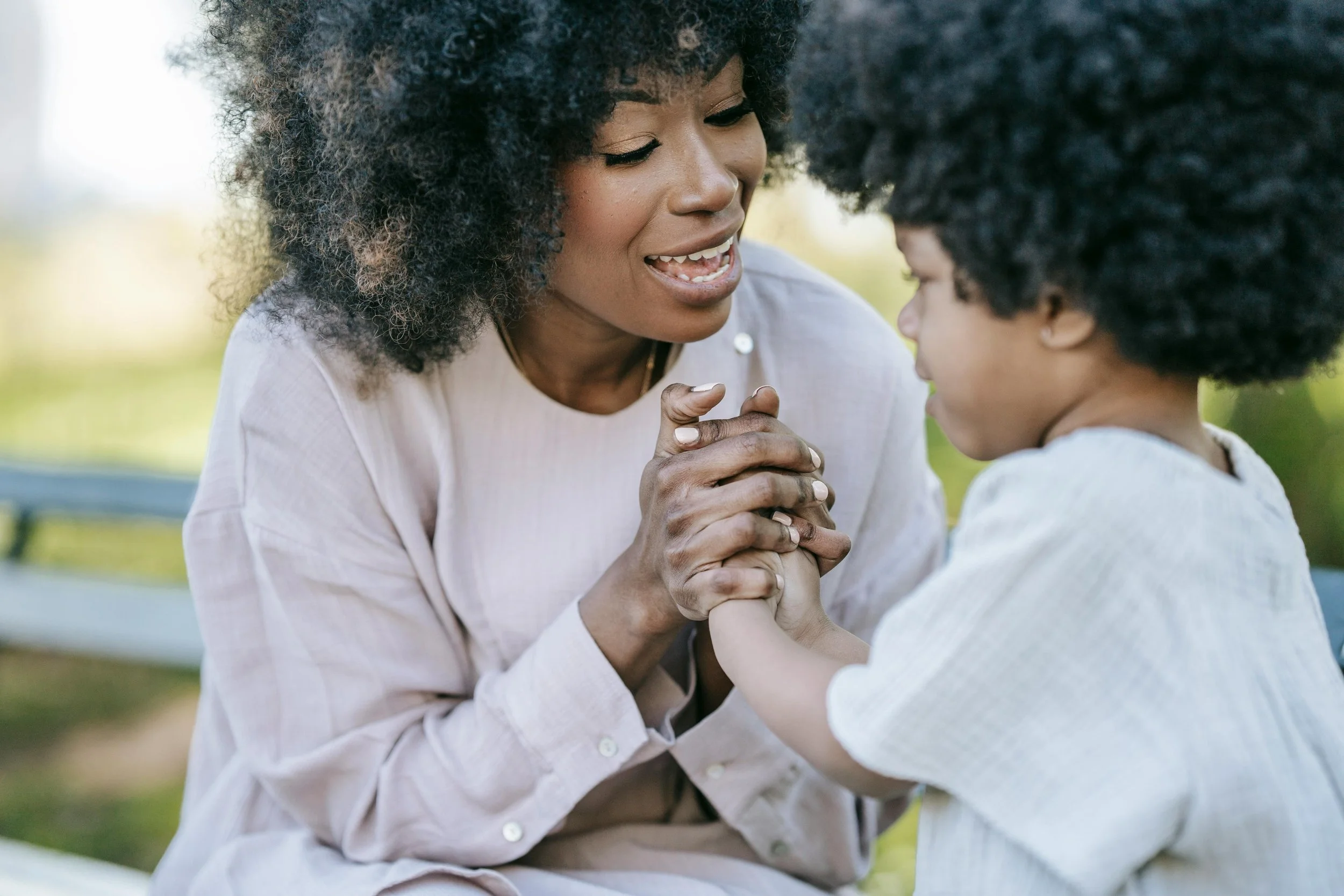 A woman and a young girl with curly hair holding hands and smiling, outdoors in a park.
