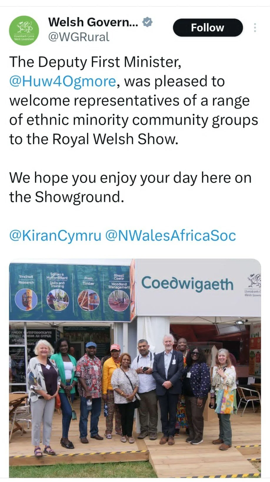 Group of diverse people standing outside a Welsh Government booth at a cultural event. The booth displays signs for Coedwig, and the background shows a blue sign with images related to forestry and training.