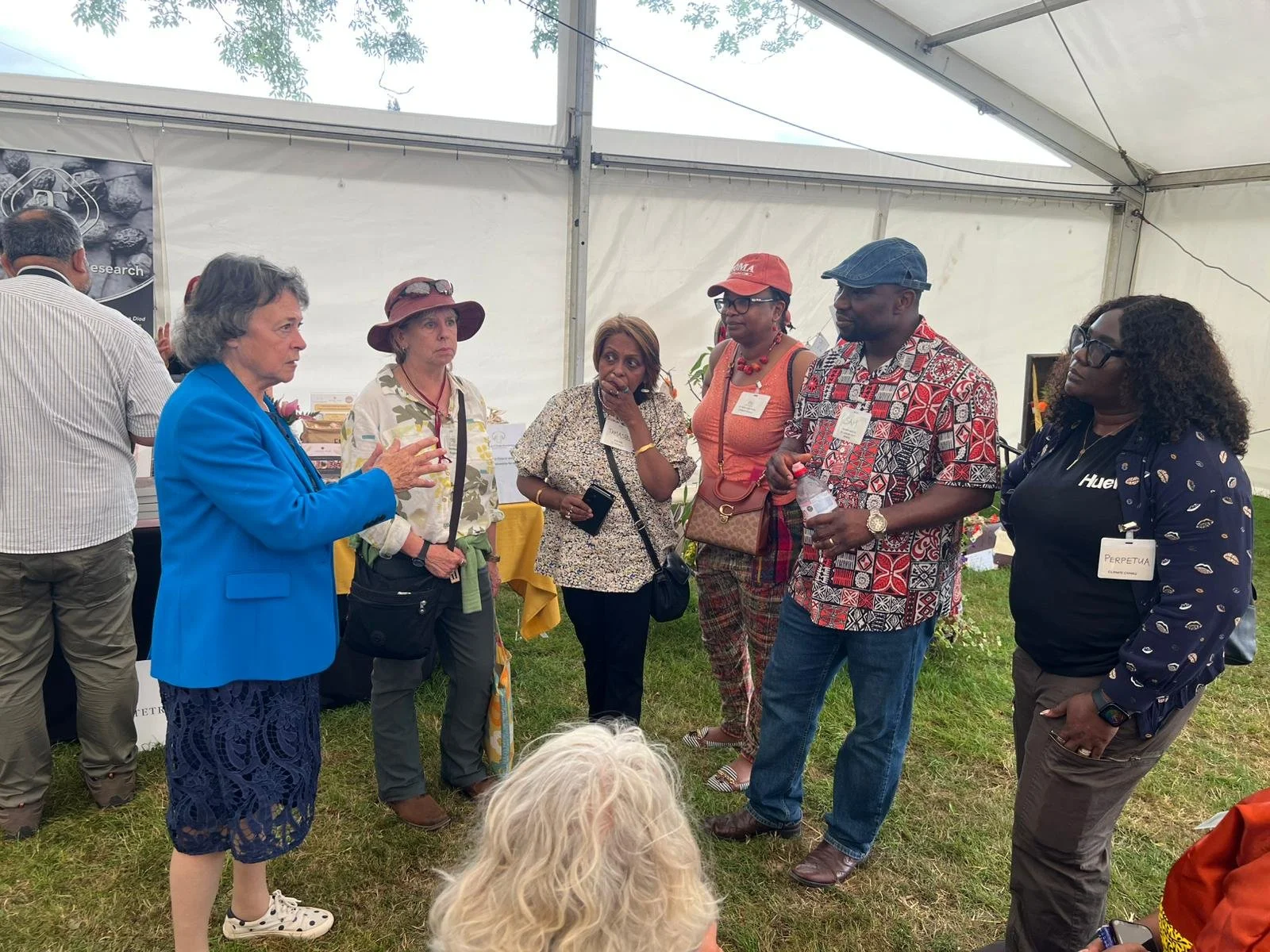 A group of six people standing in a semi-circle inside a white event tent, engaging in conversation. There are name tags on some individuals, and a woman in a blue blazer appears to be speaking to the group. The scene suggests a professional or networking event outdoors.