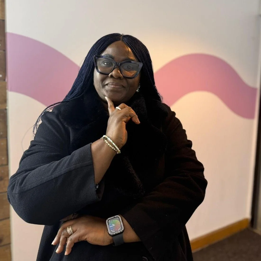 A woman with long black hair, glasses, and jewelry, wearing a black coat, standing in front of a abstract pink and white wall.