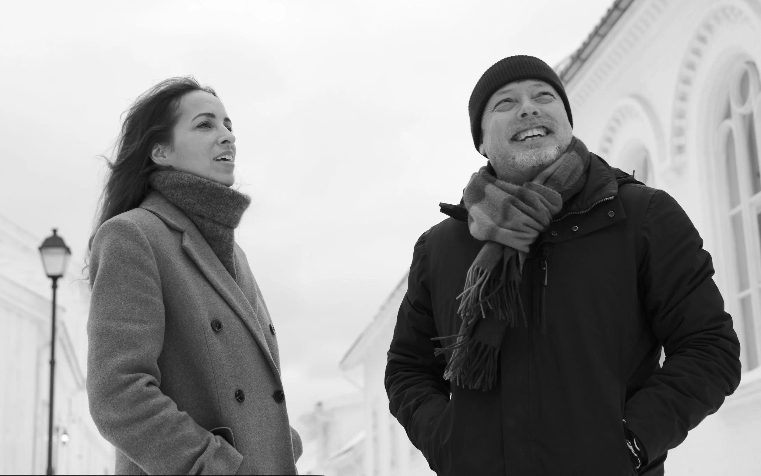 A black and white photo of a woman and a man standing outside in winter clothing. The woman is wearing a coat and a turtleneck, looking to her right, while the man is smiling, looking upwards, and wearing a beanie, scarf, and jacket.