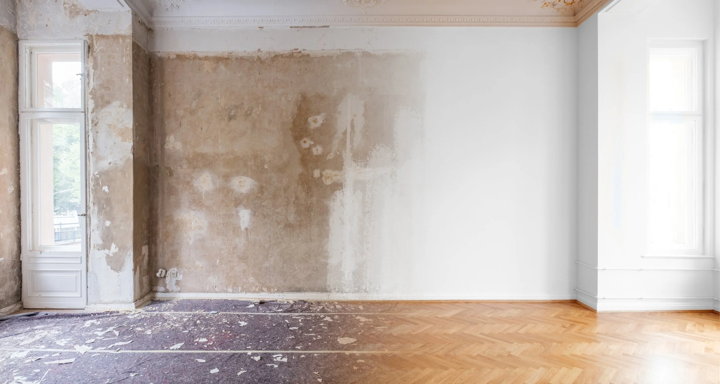 A room with one wall stripped of wallpaper and paint, showing exposed plaster and drywall on the left side. The floor on the left is unfinished with debris, while the right side features a finished wooden parquet floor. Two windows are present: one on the left with a view outside, and a larger window on the right, both letting in natural light.