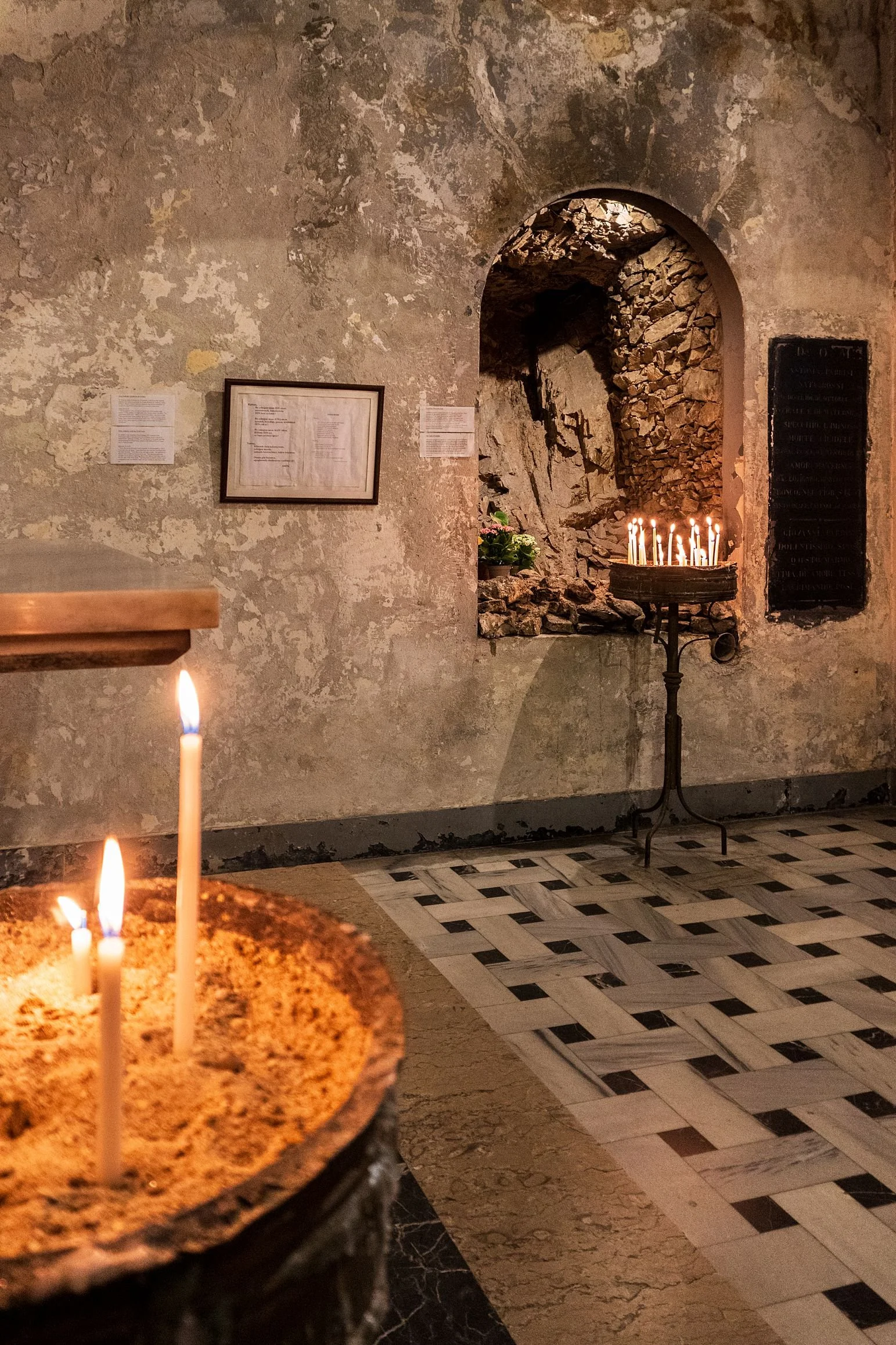 Inside a church or chapel with candles lit, weathered walls, a framed document, and a small shrine with candles and flowers.