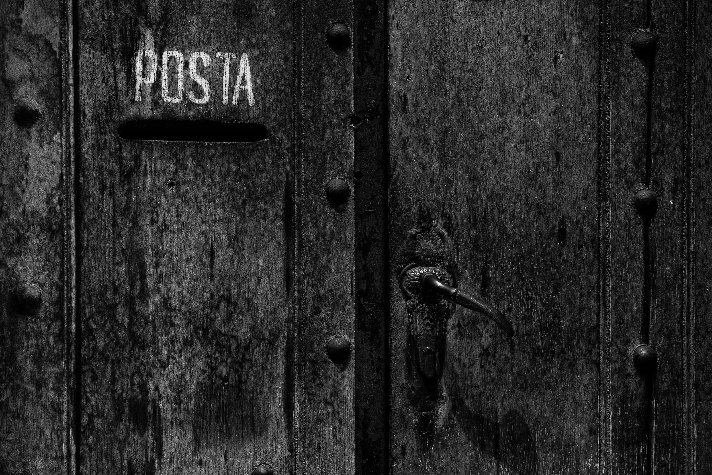 Black wooden door with a vintage door handle, rivets, and a mail slot labeled "POSTA".