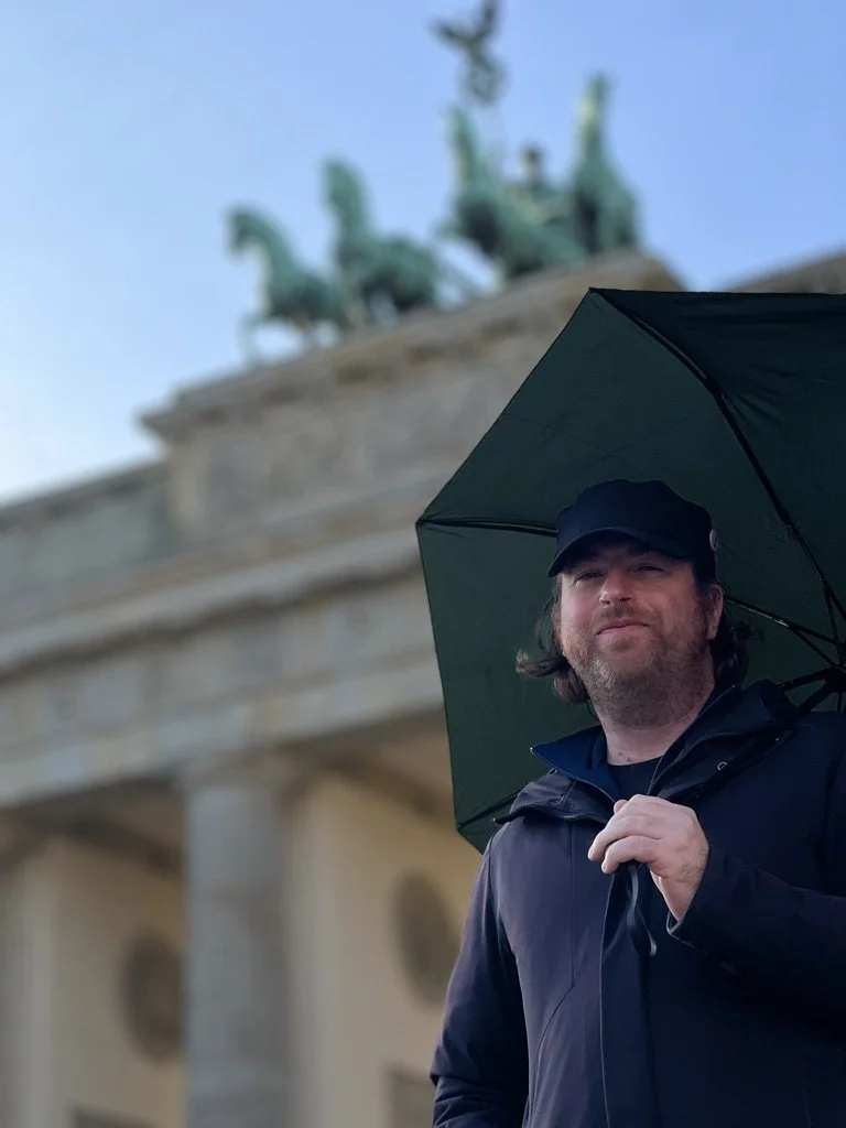 A man holding a black umbrella in front of the Brandenburg Gate in Berlin, with the Quadriga sculpture visible on top.