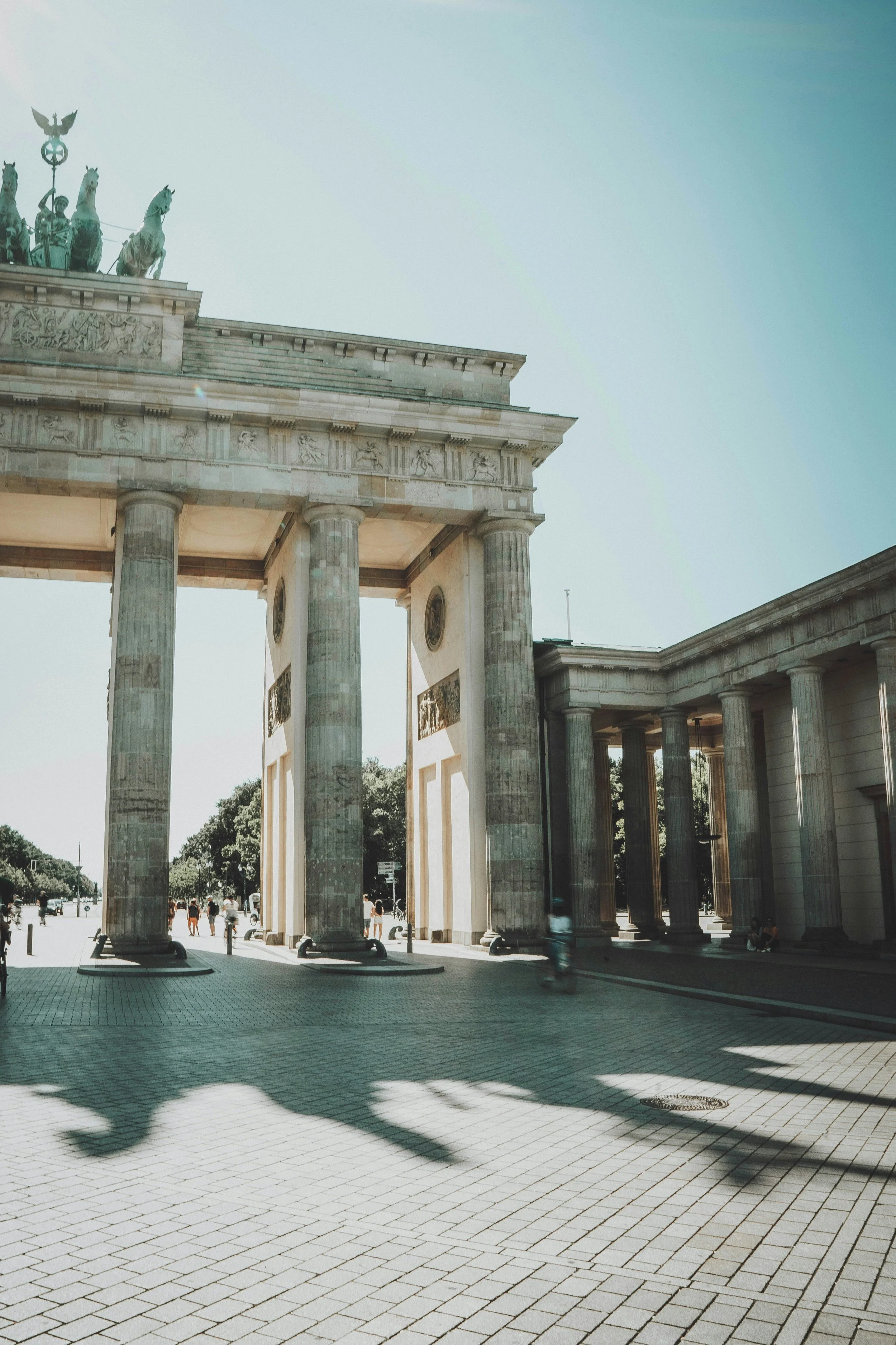 The Brandenburg Gate, an 18th-century neoclassical monument in Berlin, Germany, with large columns casting shadows on the paved ground.