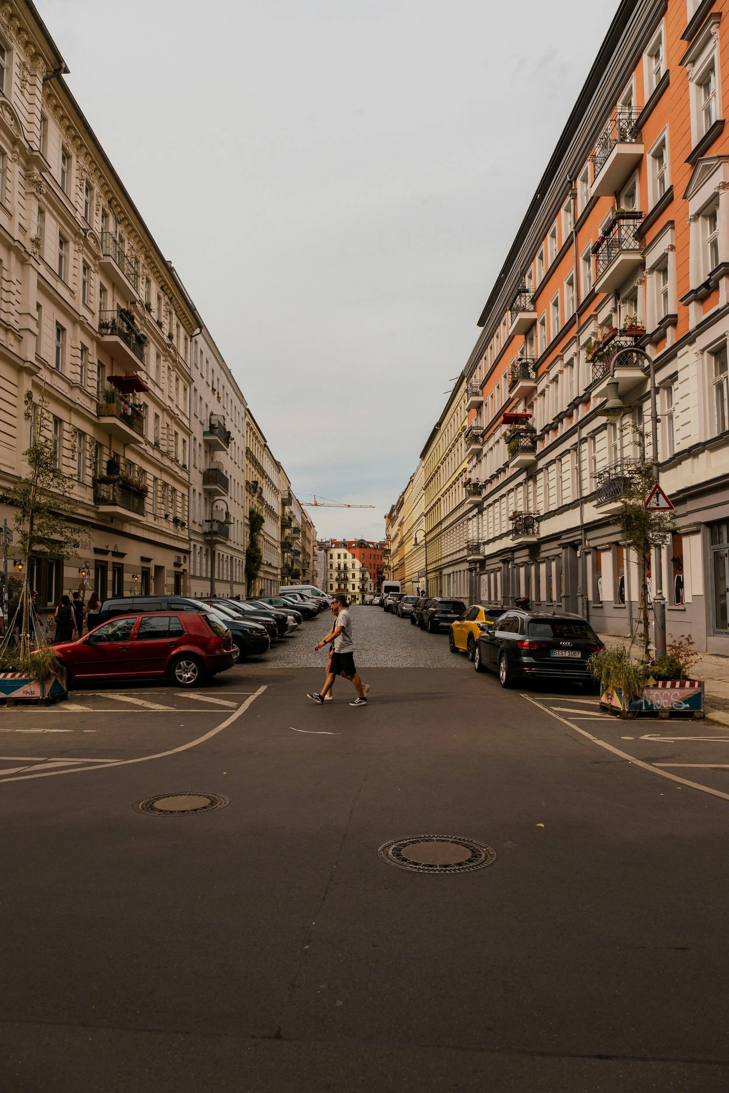 A man walking across a city street with multistory buildings and parked cars lining both sides, under an overcast sky.