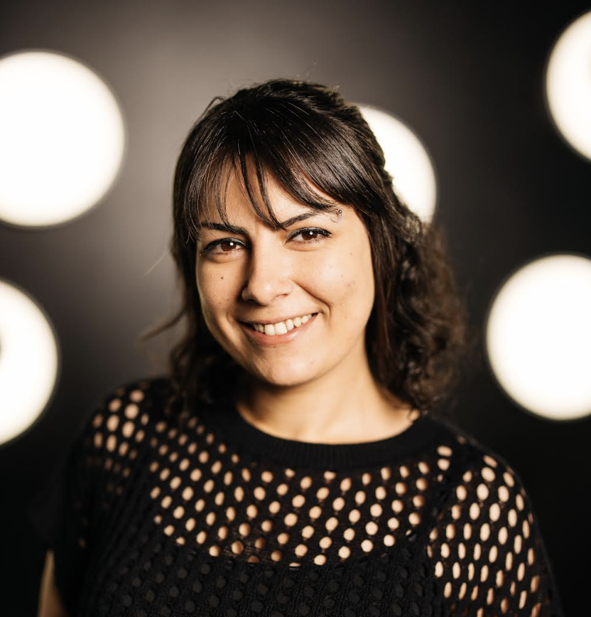 A woman with dark hair, smiling and wearing a black mesh top, standing in front of a dark background with bright circular lights.