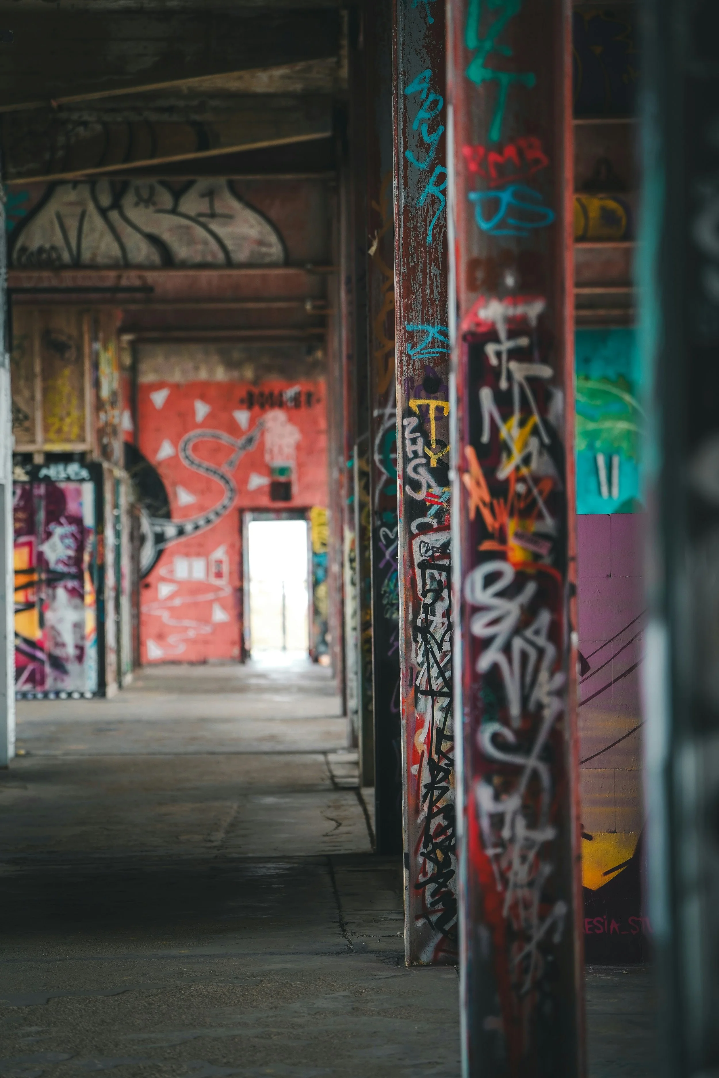 View of an abandoned building interior with graffiti-covered walls, metal beams, and an open door at the end of the corridor leading to the outside.