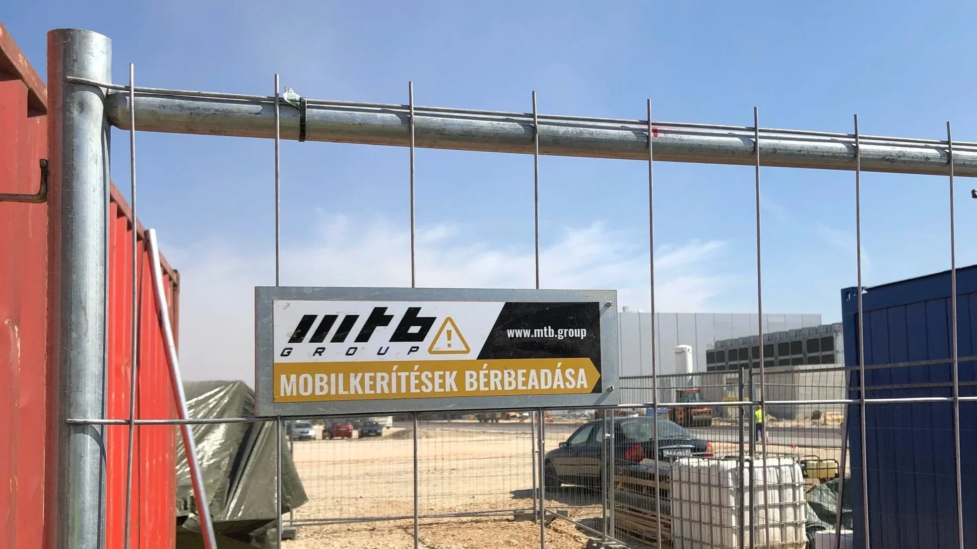 Kerítés, céges felirattal amin az olvasható magyarul, hogy "Mobilkerítés bérbe", egy építkezésen, napos, enyhén felhős égbolttal, parkoló autók, építkezés, konténerek a háttérben.