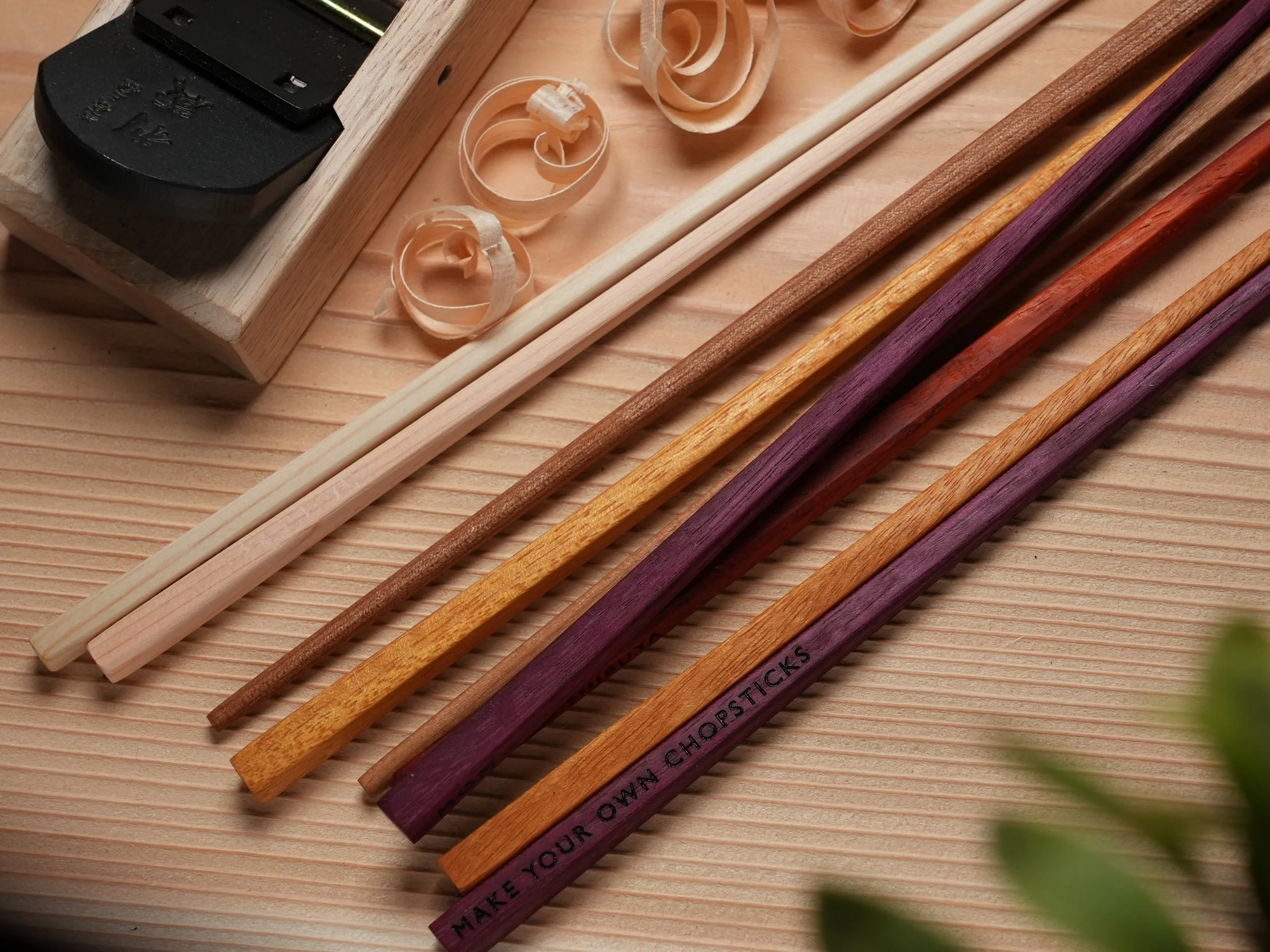 Assorted colored and natural wood Chopsticks and shavings on a light wood surface. a traditional japanese handplane known as a kanna is in the corner. Maple, Purple-heart wood, Ceder, Hinoki Cypress, Karin Wood. Wooden shavings.
