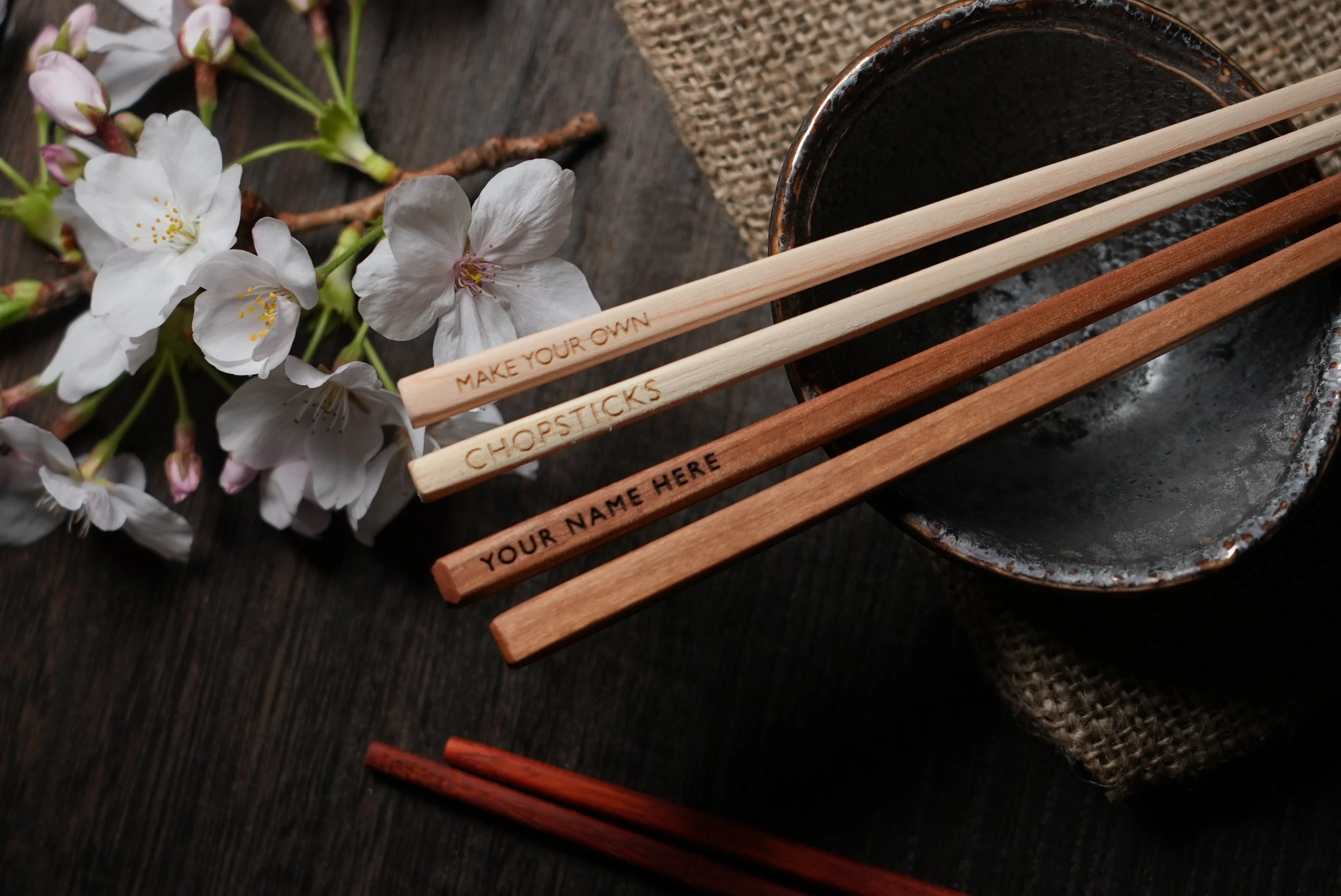 White cherry blossom flowers (Sakura) and wooden chopsticks on a dark wooden surface near a nice ceramic black bowl. the chopsticks have a custom engraving saying make your own chopsticks, your name here. they are masterfully handmade and finished.
