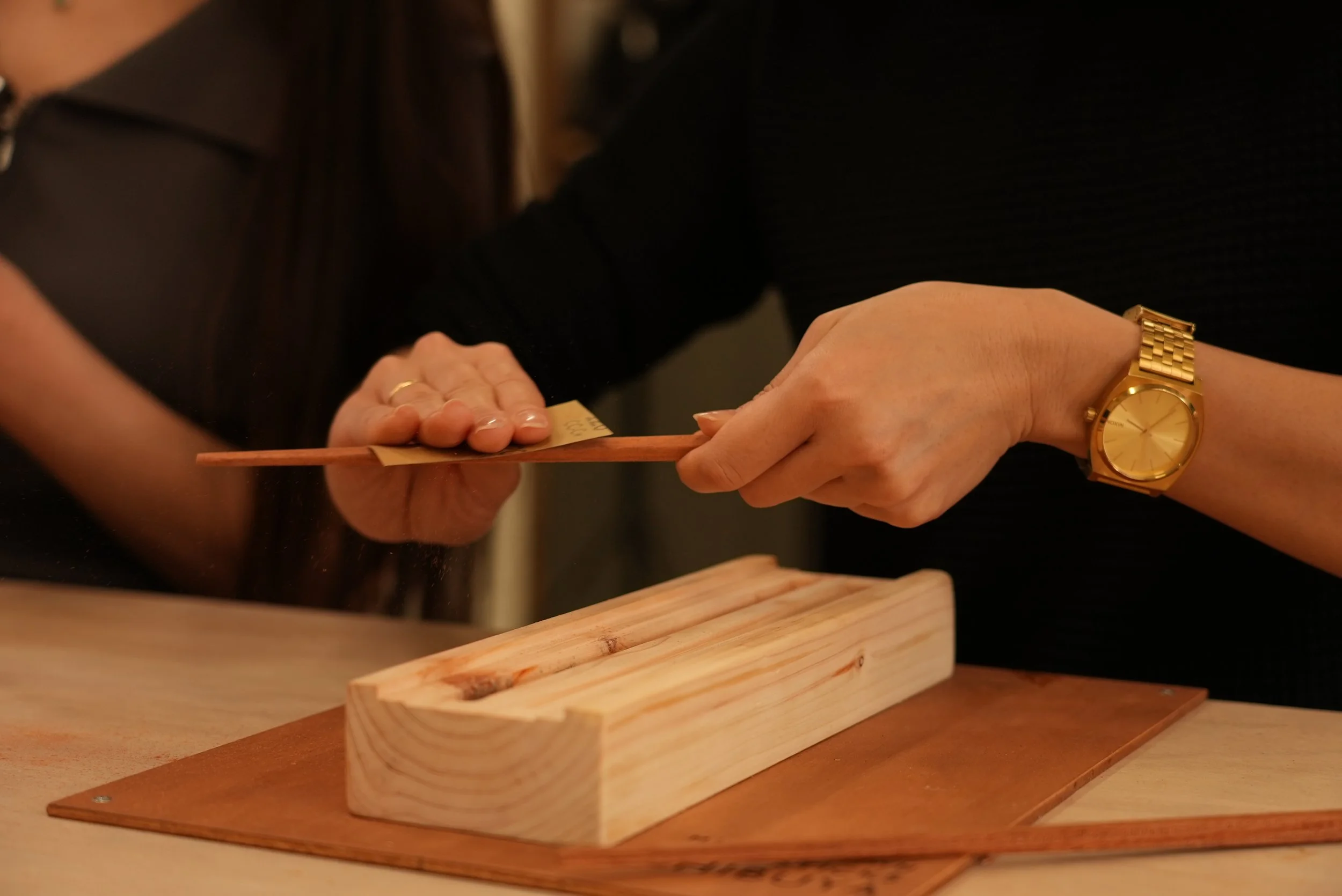 A close up of a Japanese girls hand sanding her own chopstick over a custom made chopstick carving jig. she has a golden ring and watch on. she is perfecting her chopstick.