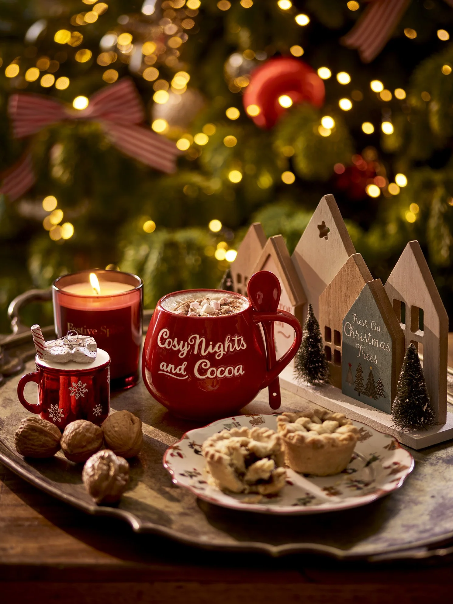 A cozy Christmas scene featuring a tray with cookies, walnuts, and mugs of hot cocoa, surrounded by candles and decorated with a Christmas village, lit Christmas tree, and ornaments in the background.