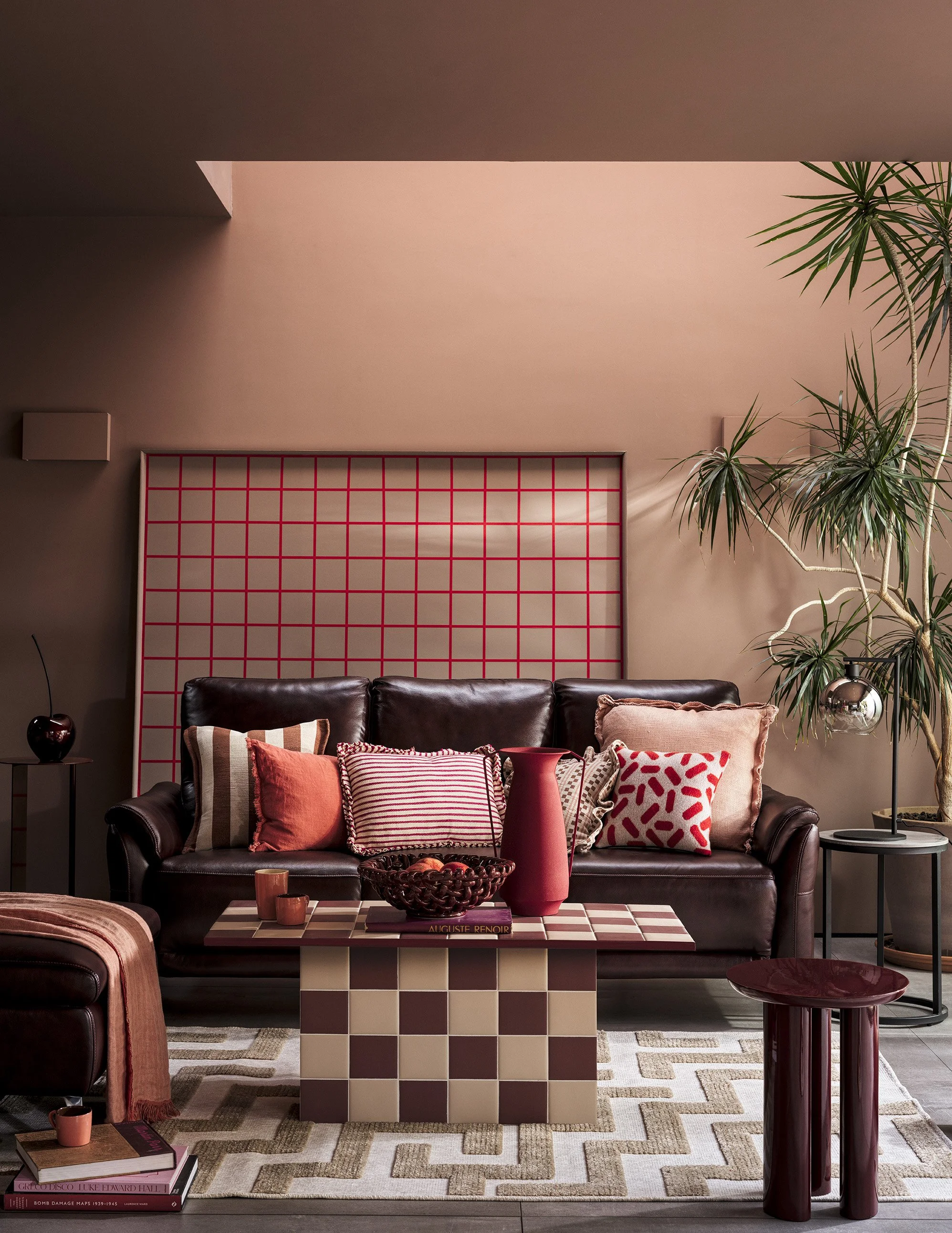 Living room with a dark leather sofa decorated with patterned cushions, a modern checkered coffee table, and green houseplants. The room features a pink wall with a grid pattern and a zigzag-patterned rug on the floor.