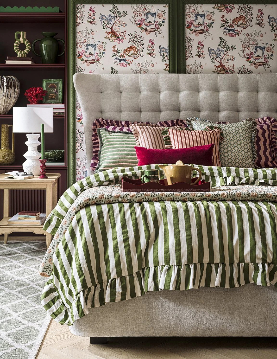 A cozy bedroom with a bed covered in green and white striped bedding, topped with multiple patterned pillows in red, pink, green, and blue. The bed has a large, light gray, tufted headboard. Behind the bed, there's wallpaper with a print of animals l