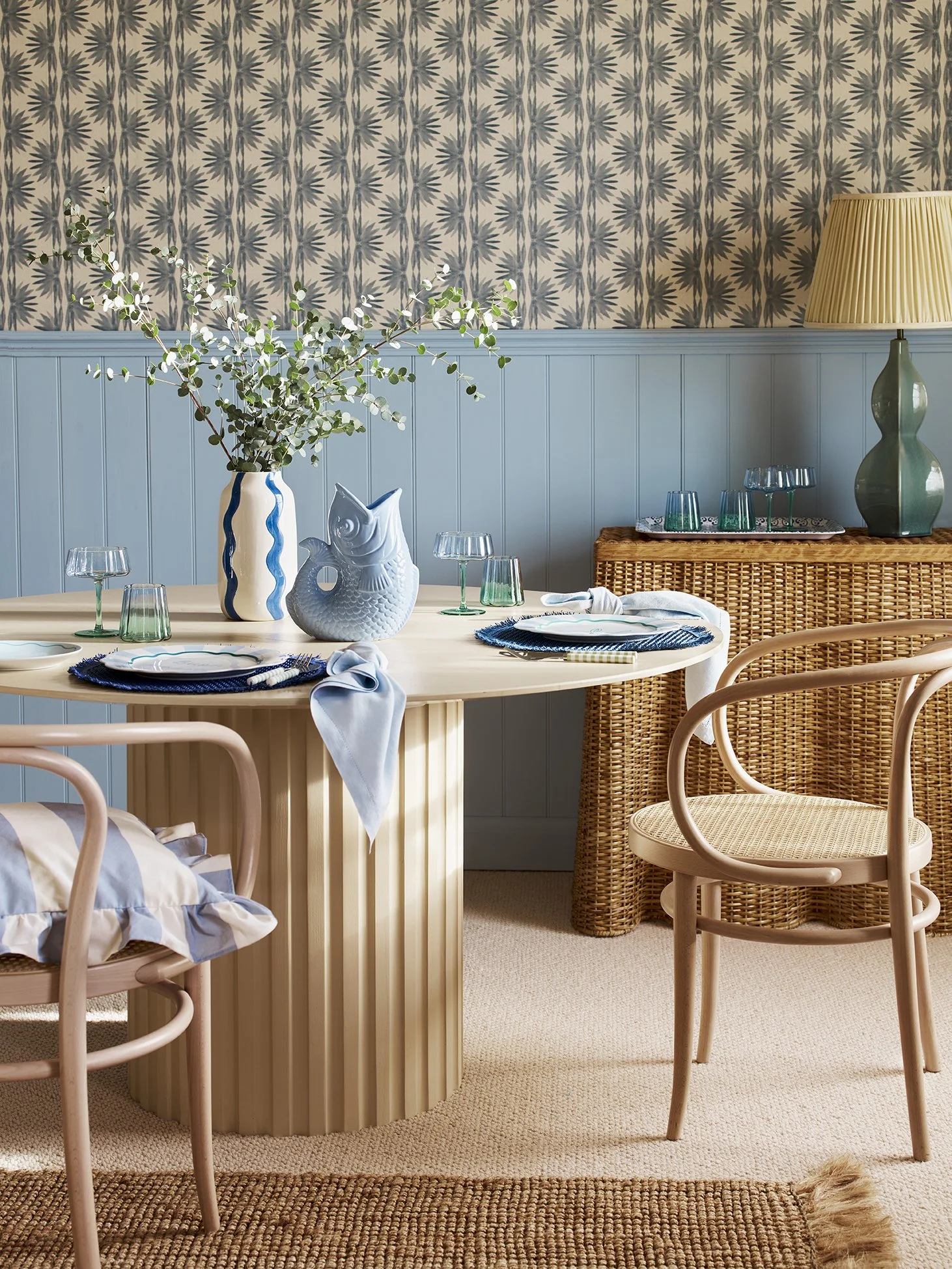 A cozy dining room with a round wooden table set with place settings, a white fish-shaped vase, and a tall blue and white striped vase with greenery. There are pastel pink chairs, a rattan sideboard, a green table lamp, and a patterned wallpaper back